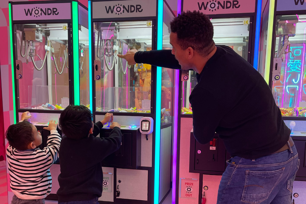 Een man begeleidt twee kinderen bij grijpmachines met kleurrijke lampjes.
