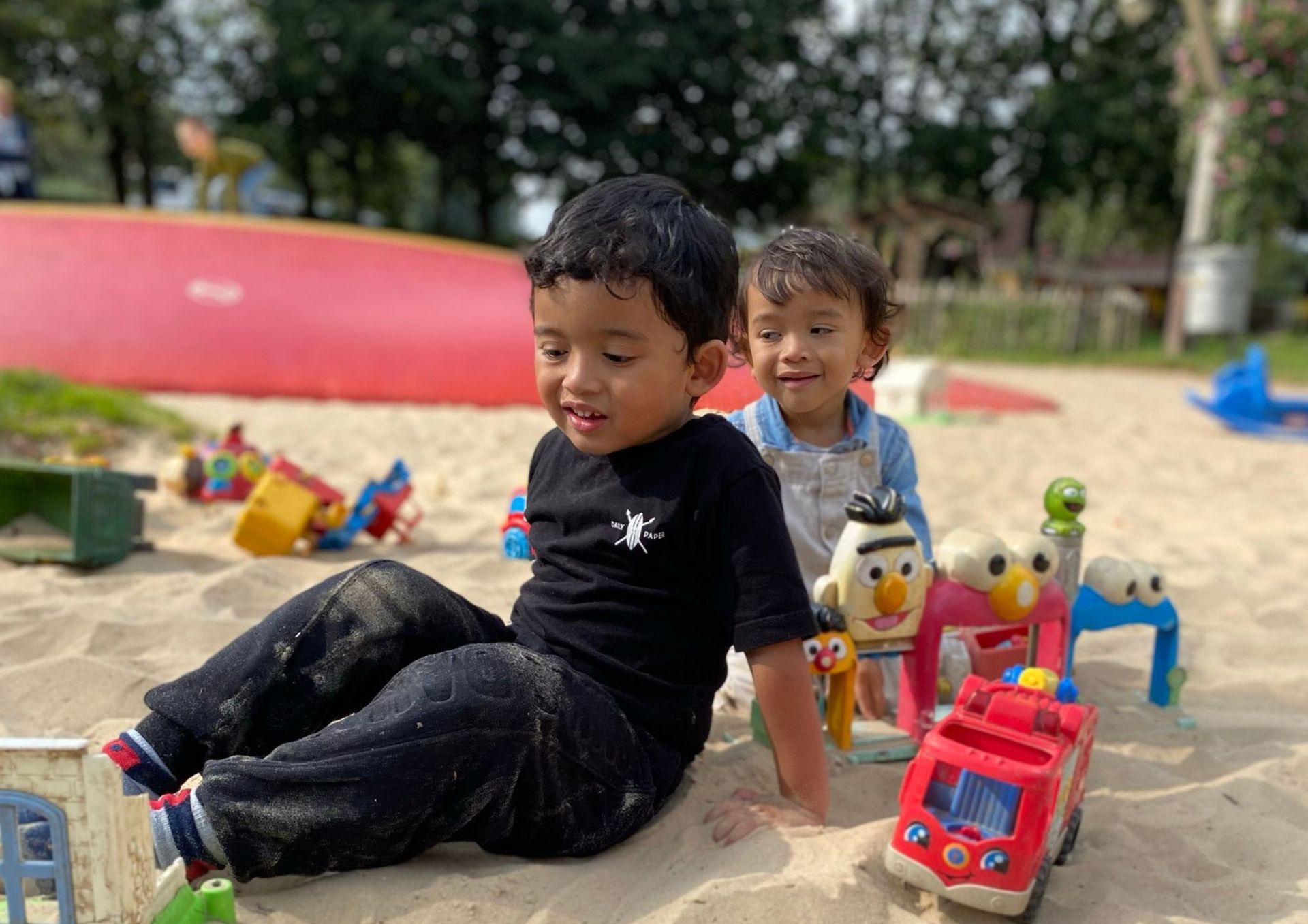 Twee kinderen spelen met speelgoed in een zandbak. De ene heeft een zwart shirt aan, de andere een lichtblauw shirt.