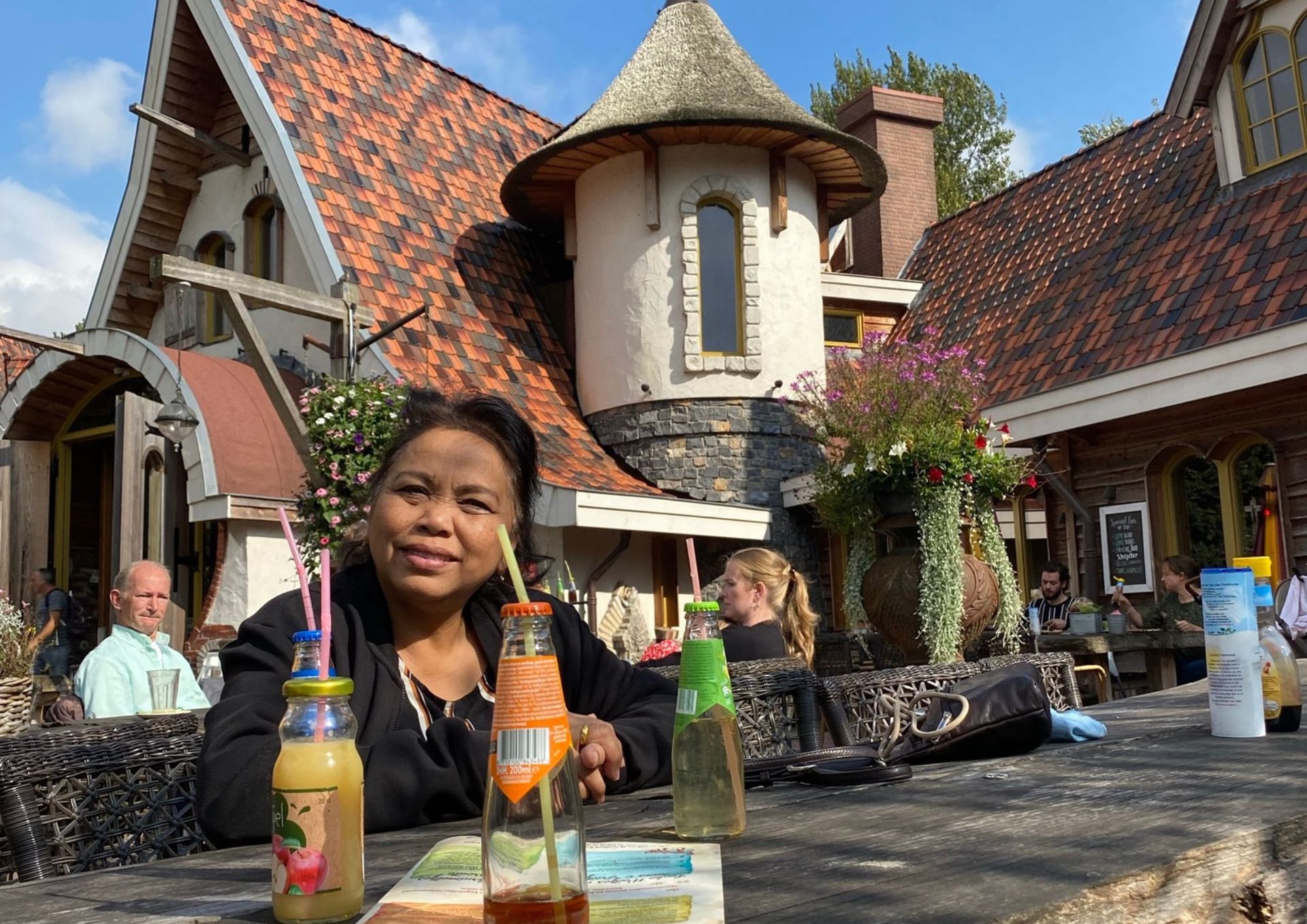 Vrouw aan een tafeltje buiten, met een drankje voor zich. Op de achtergrond een gebouw in fantasiestijl.