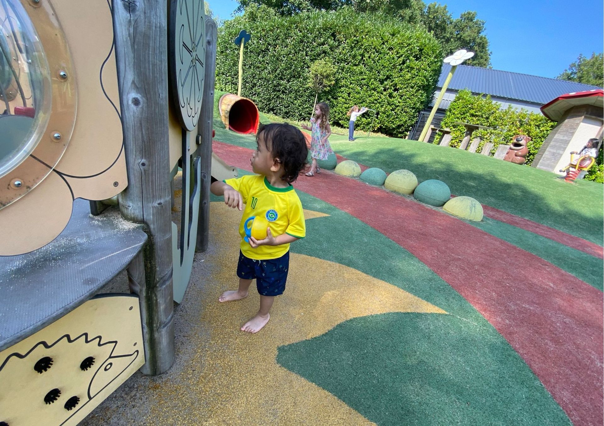 Een kind in een geel shirt speelt op een kleurrijke speeltuin met houten speeltoestellen en een rood-groen pad.