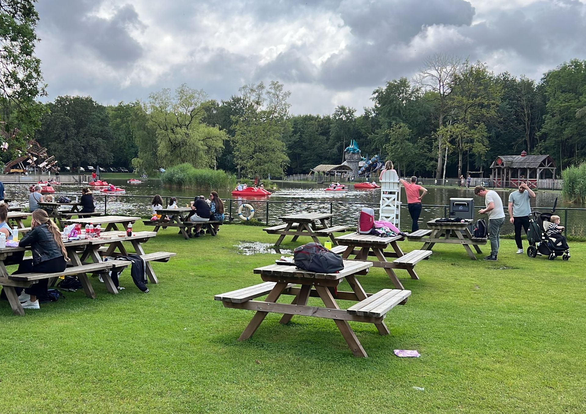 Picknicktafels op een grasveld naast een vijver. Mensen zitten aan tafels, sommigen staan, anderen hebben een kinderwagen. Op de achtergrond zijn bomen en een bewolkte hemel te zien.