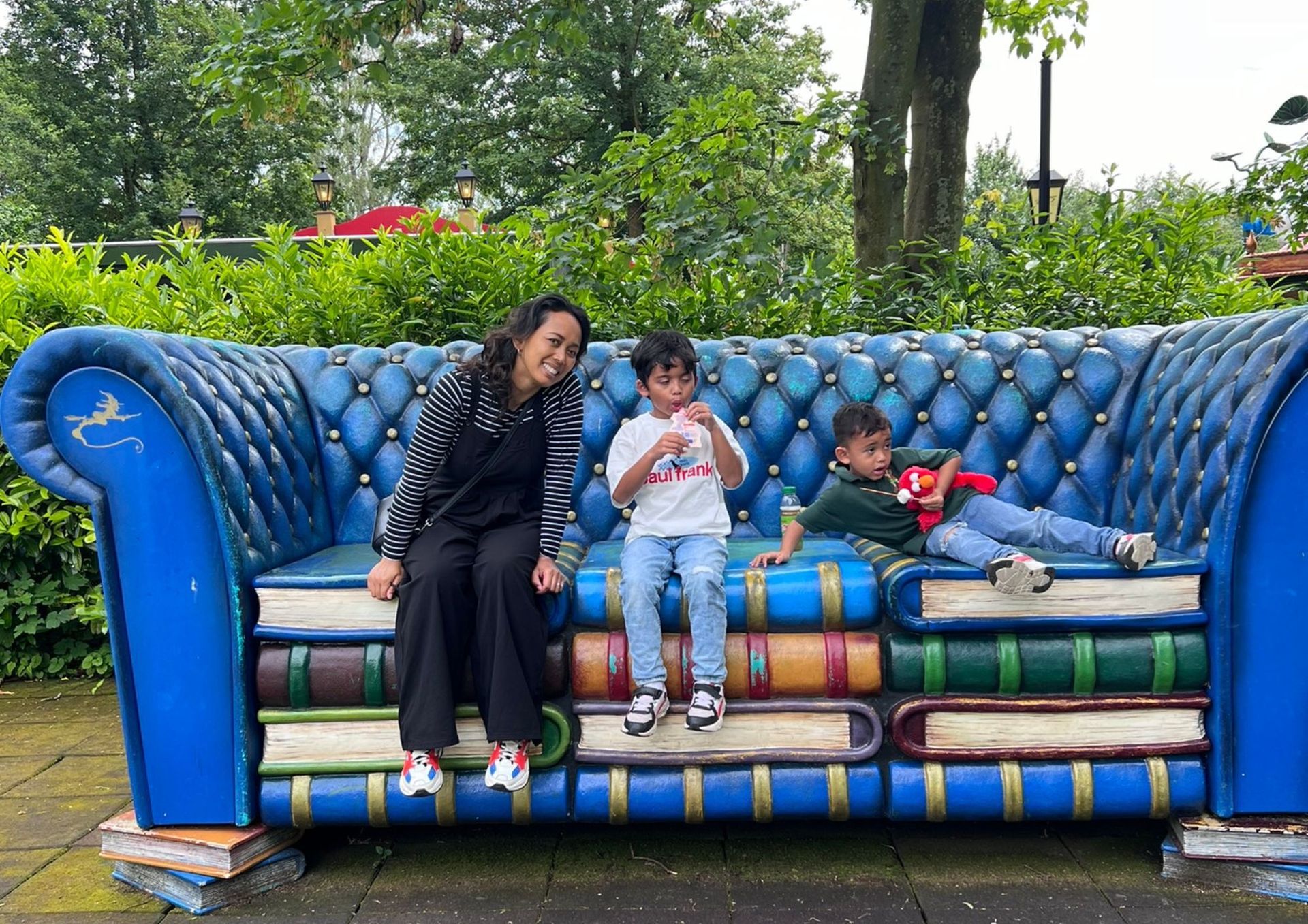 Een vrouw en twee kinderen zitten op een blauwe bank in de vorm van boeken in een park.