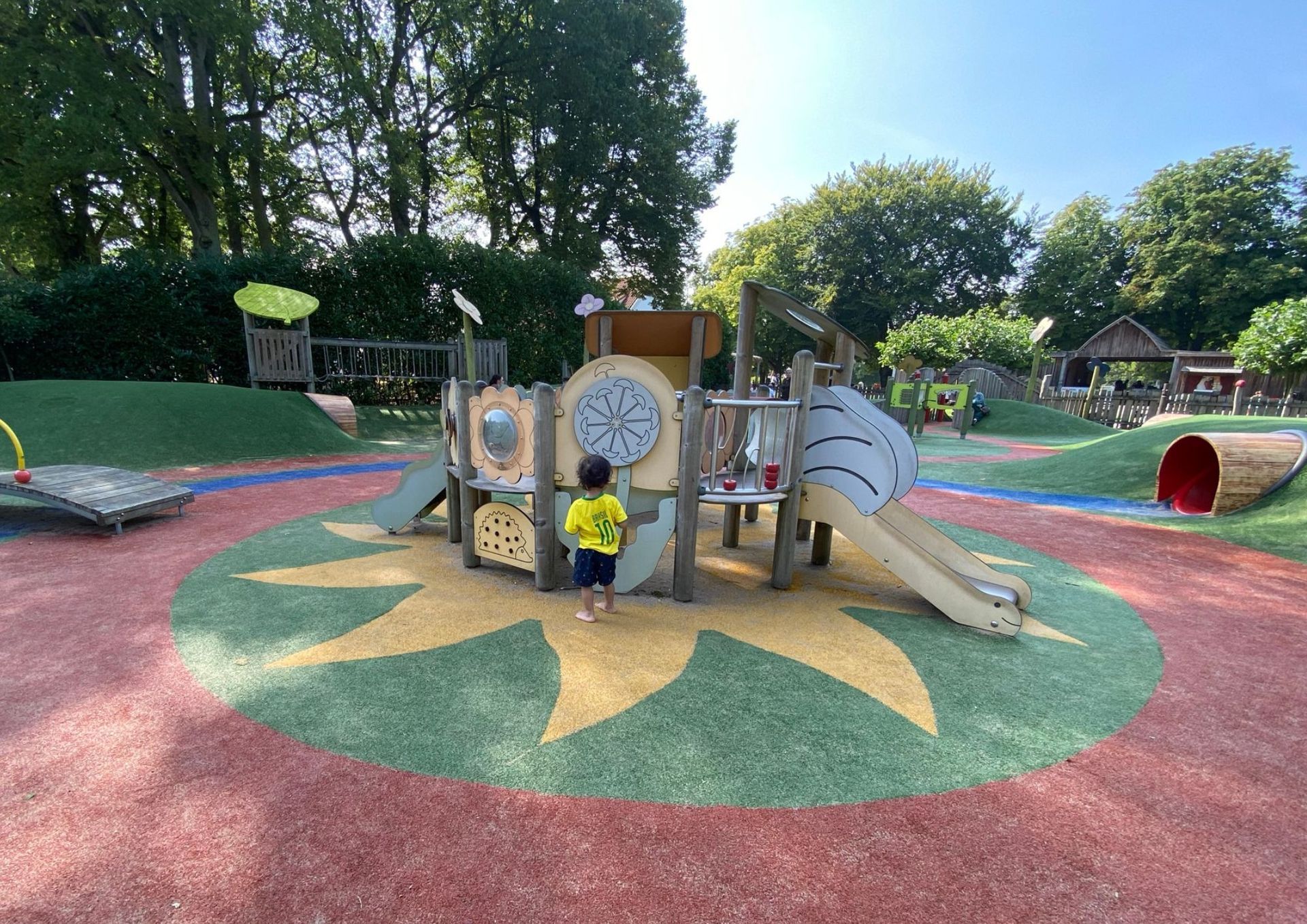 Kind op een kleurrijke speeltuin met glijbanen, klimtoestellen en groene heuvels.