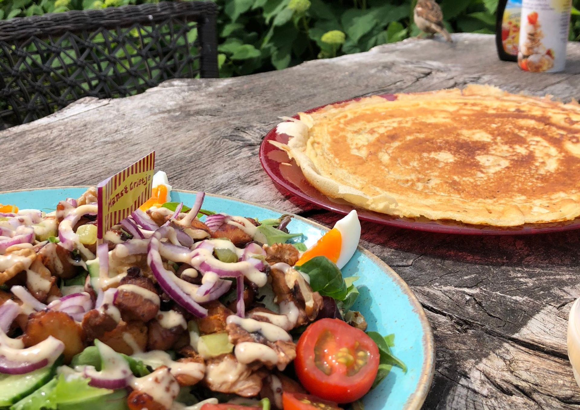 Salade met diverse toppings en een pannenkoek op een houten tafel, buiten.