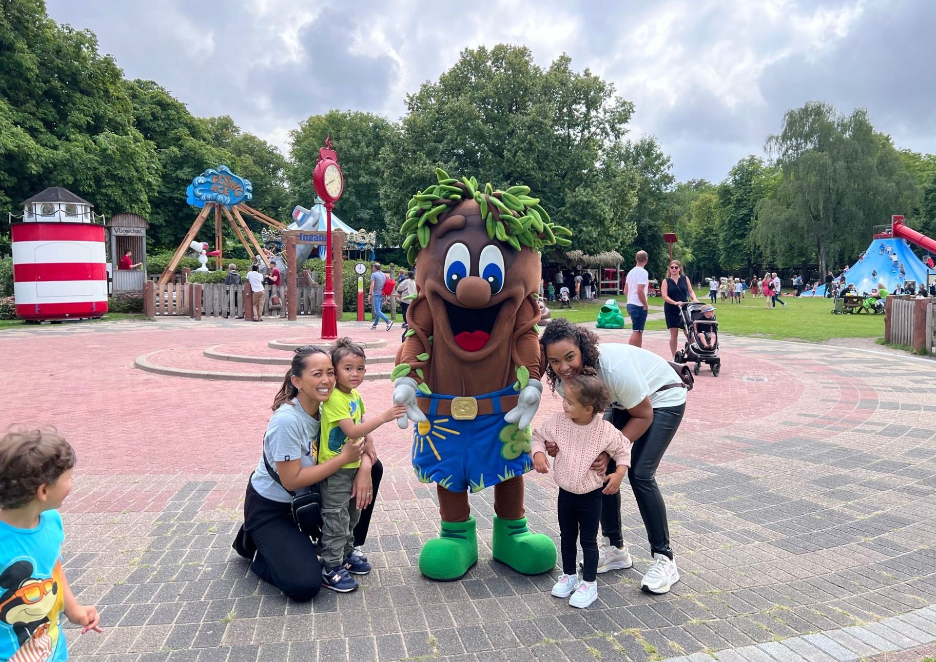 Mensen poseren met een mascotte in een openluchtpark. Op de achtergrond zijn een speeltuin en groen te zien.