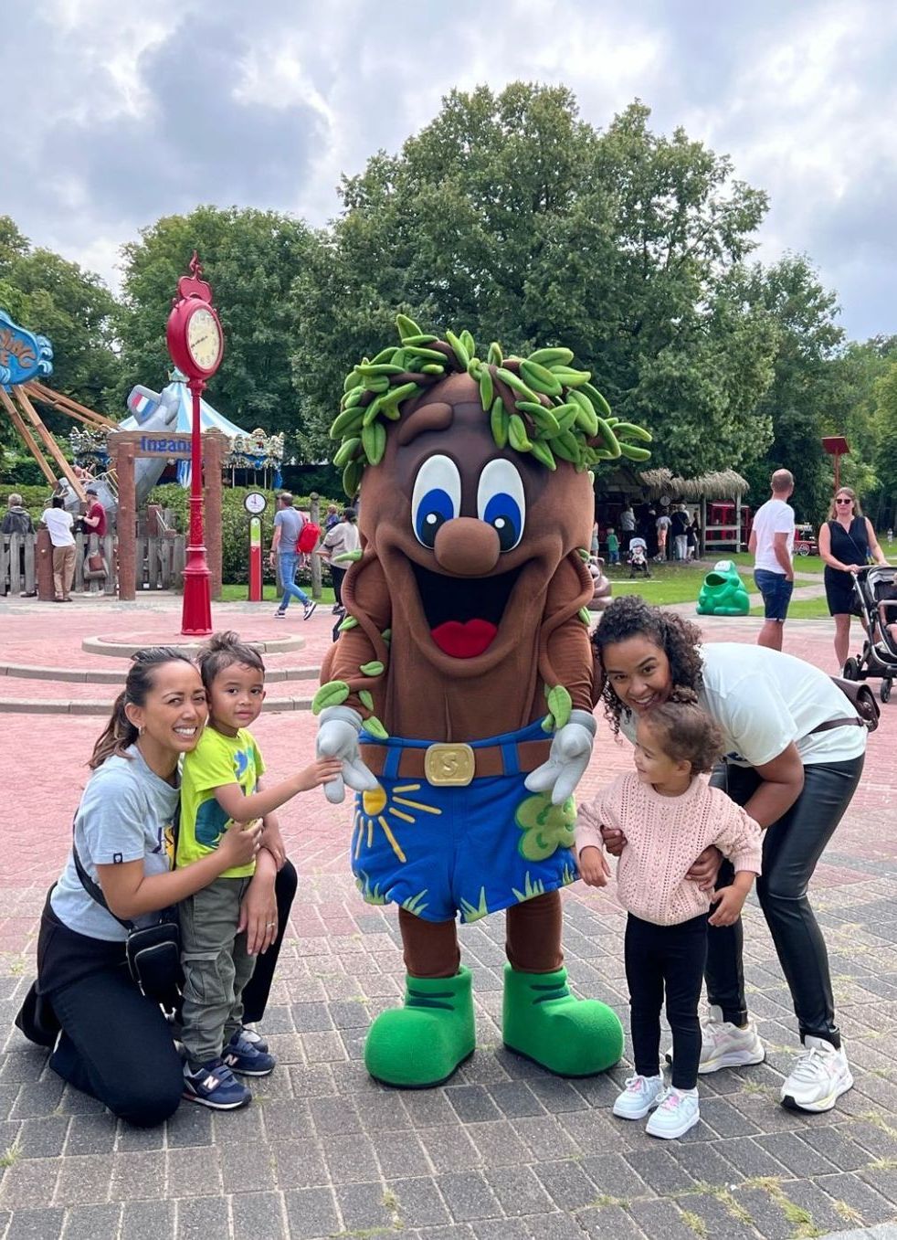 Twee volwassenen en twee kinderen poseren lachend met een parkmascotte in de buitenlucht op een zonnige dag.