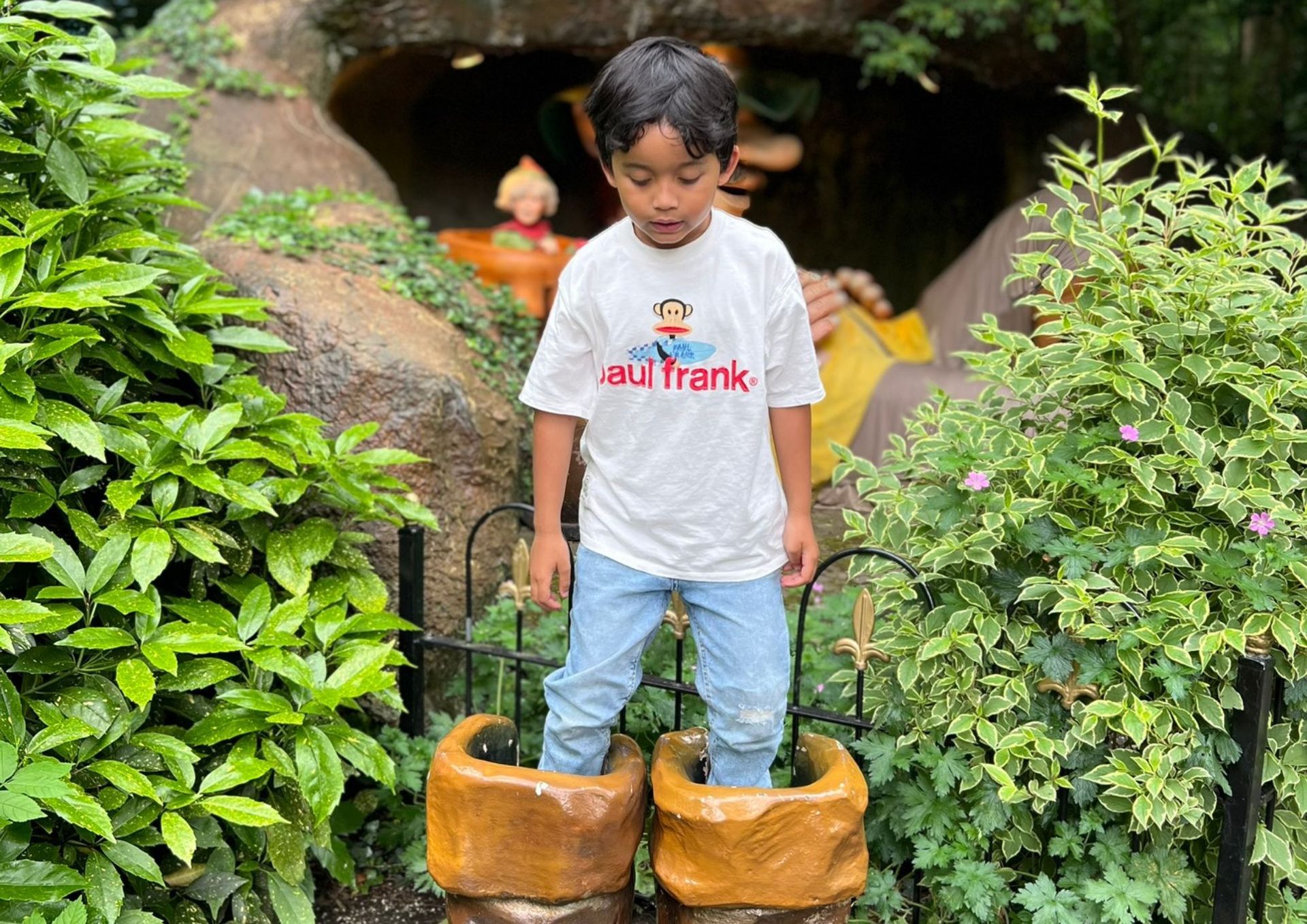 Een jongen, gekleed in een Paul Frank T-shirt en spijkerbroek, staat op houten klompen in een tuin.