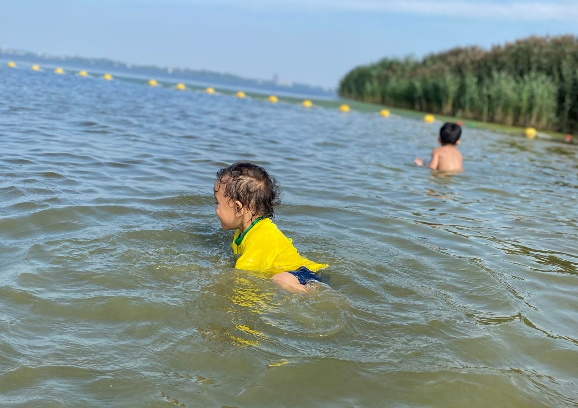 Twee peuters spelen in een meer; de ene draagt ​​een geel shirt, de andere staat wat verder naar achteren. Rustig water, zonnige dag.
