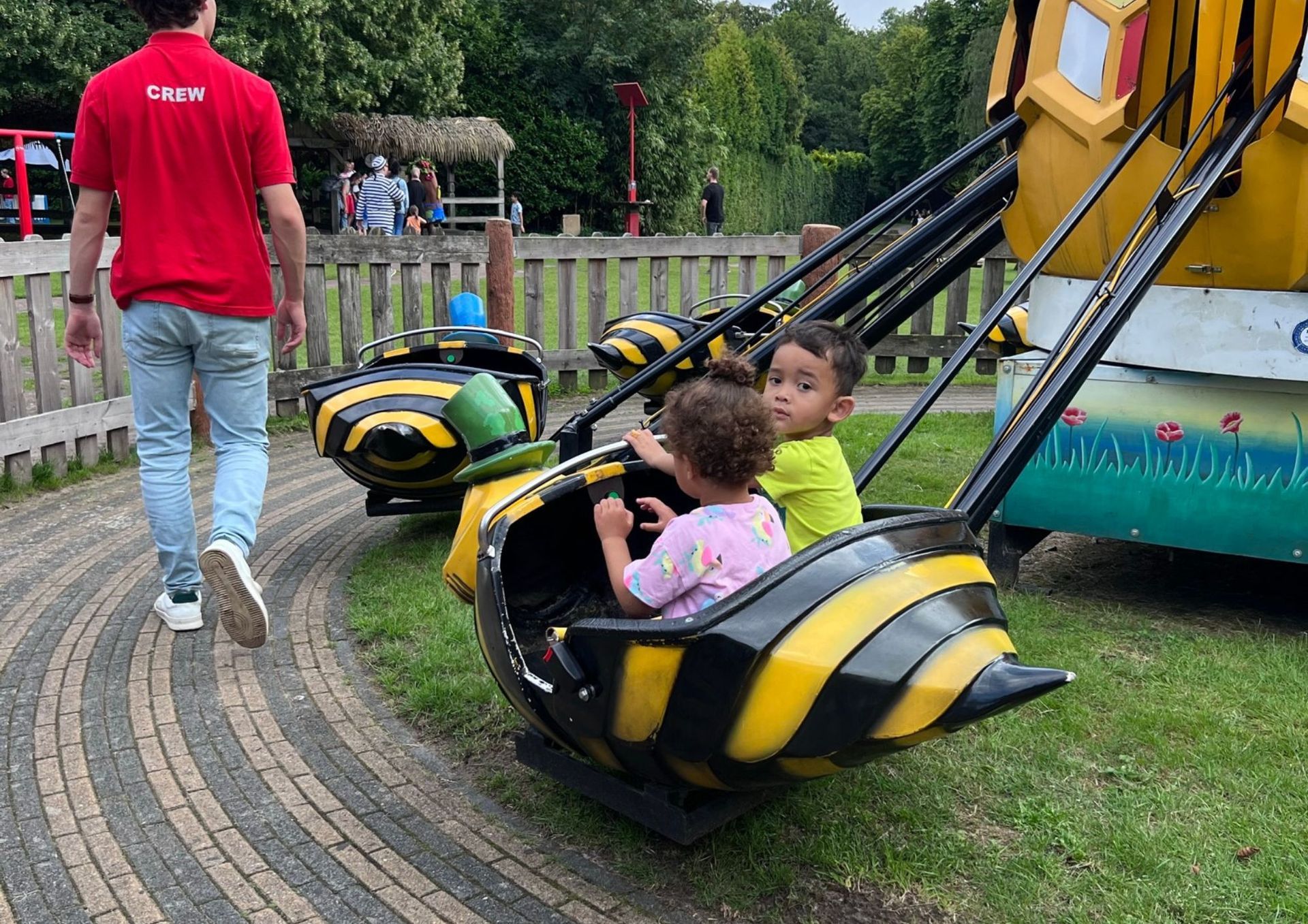 Twee kinderen zitten in een geel-zwarte attractie met een bijenthema. Een persoon in een rood shirt loopt naast de attractie.