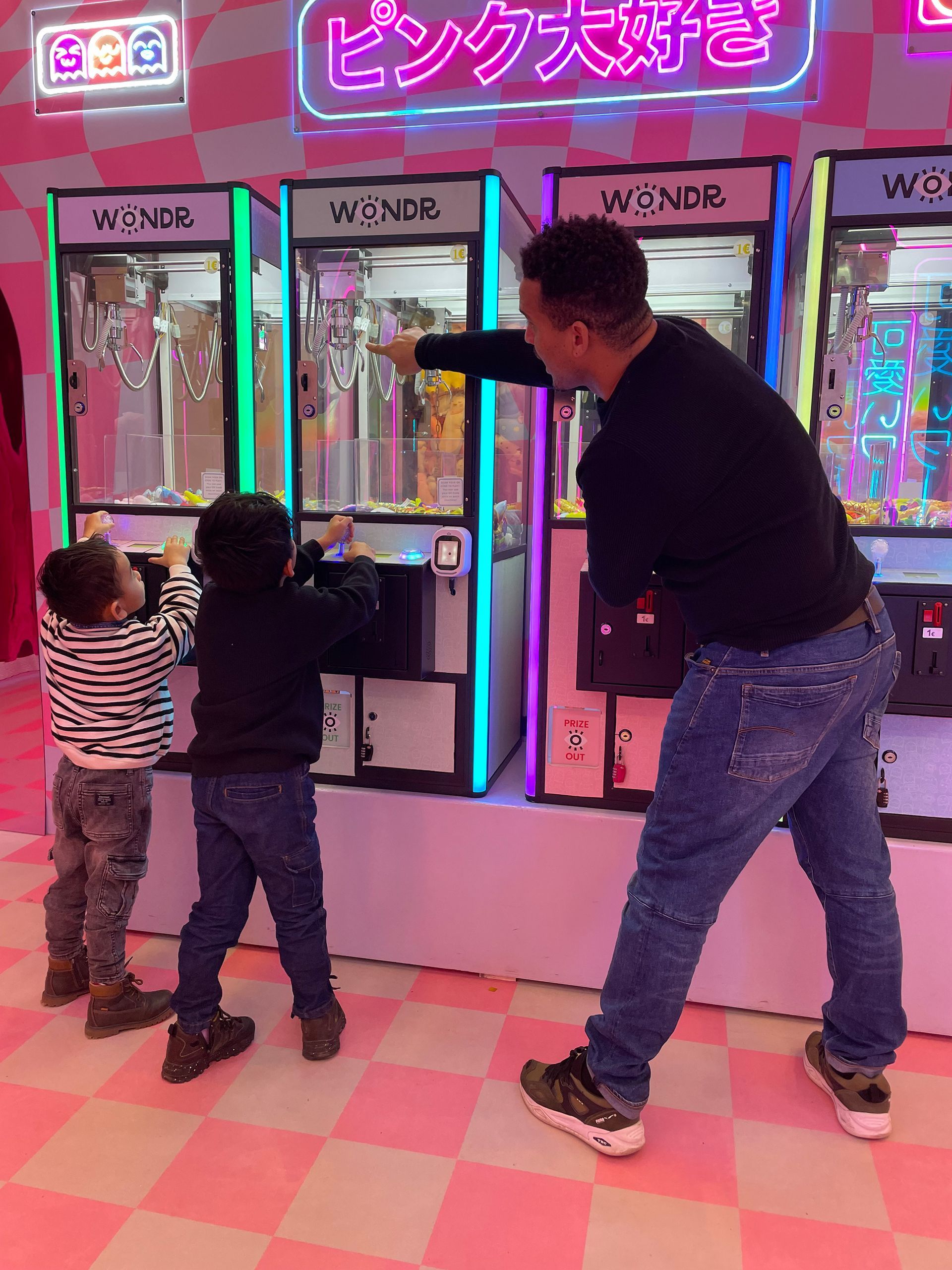 Een man en twee kinderen spelen met grijpmachines in een roze speelhal met neonverlichting.