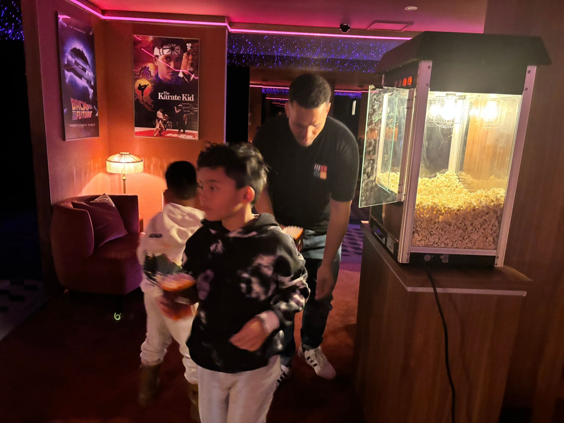 Een man en twee kinderen staan ​​bij een popcornmachine in een thuisbioscoop, die verlicht wordt door kleurrijke lampen.