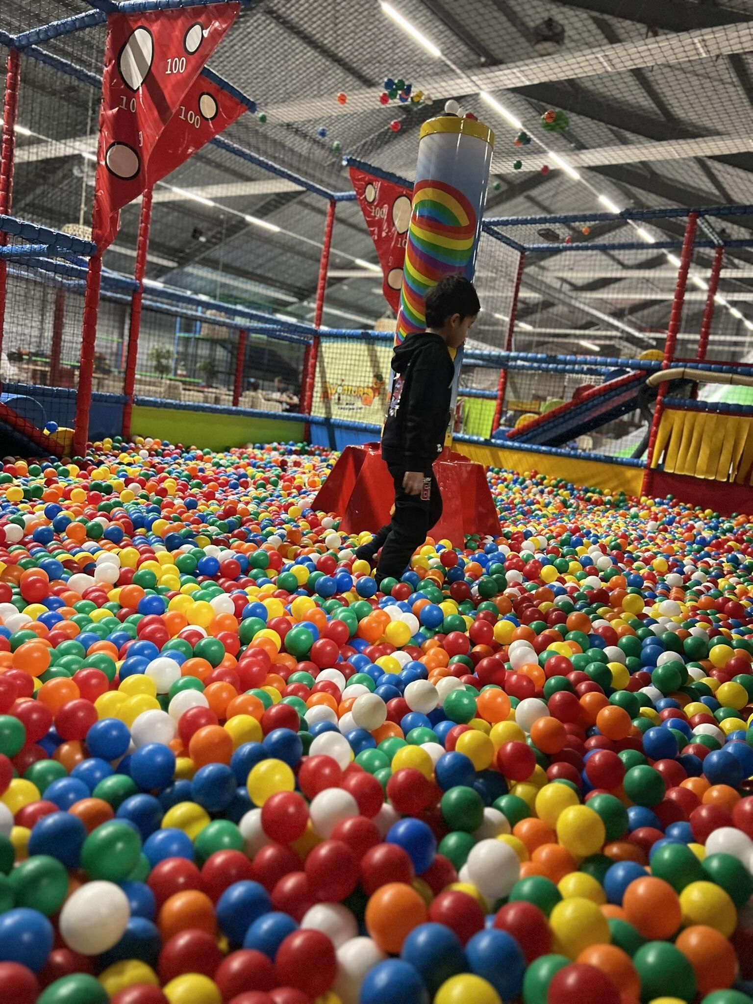Een kind springt in een kleurrijke ballenbak in een binnenspeeltuin, met zichtbare klimtoestellen.