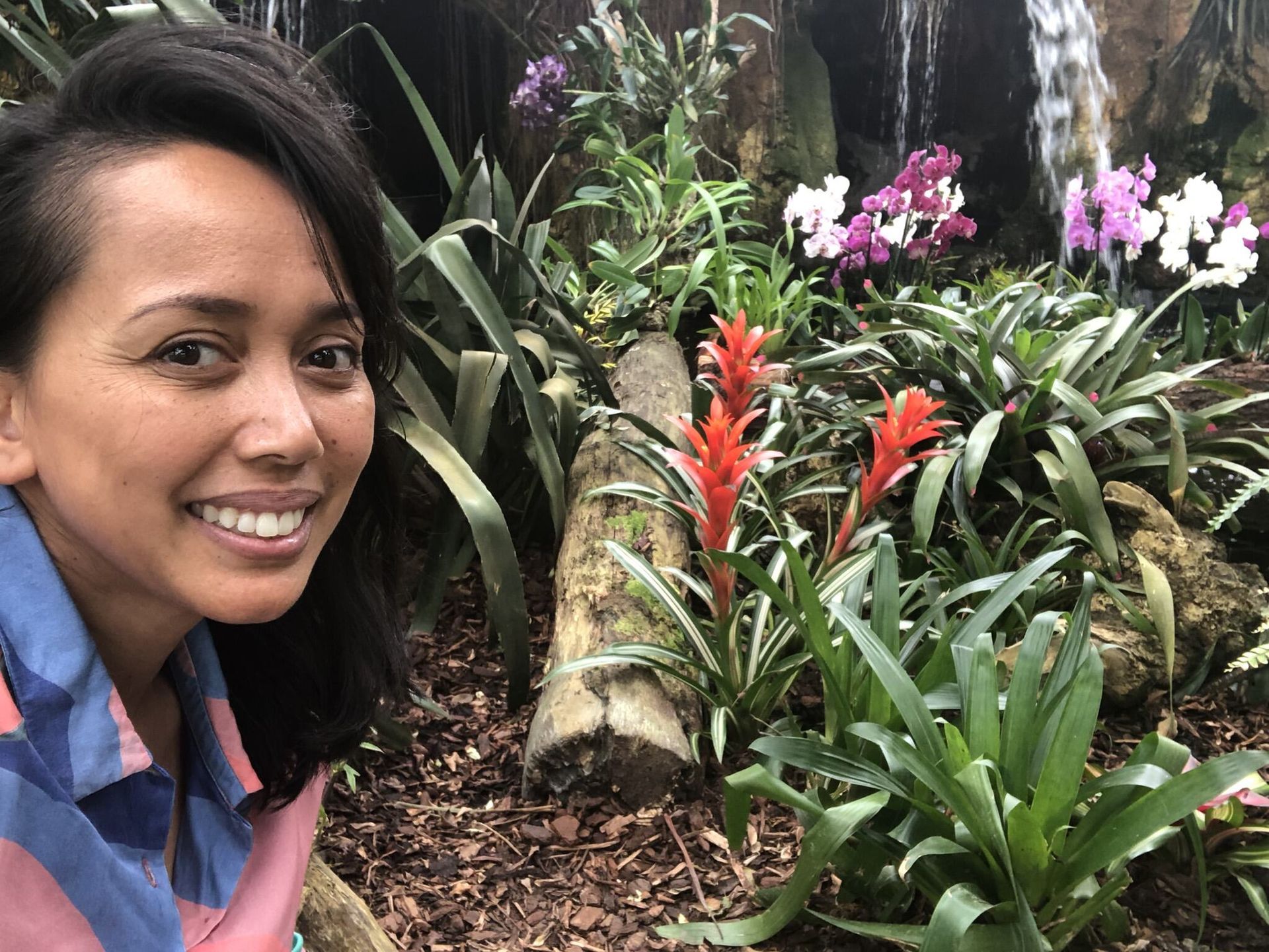 Vrouw lacht voor een tropische tuin met kleurrijke bloemen en een kleine waterval.