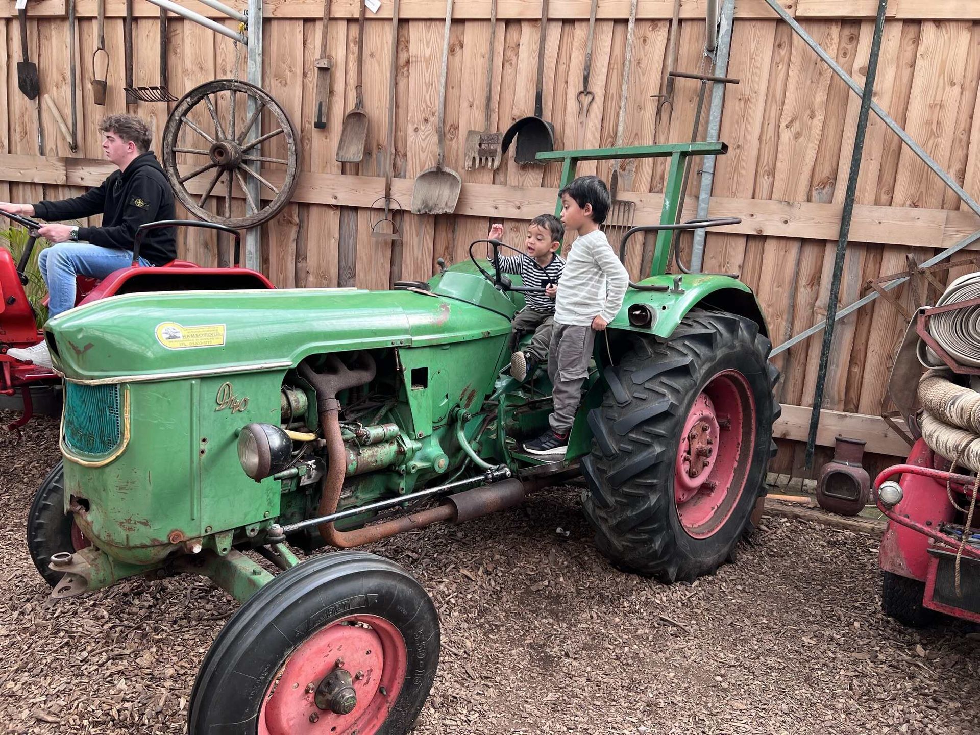 Drie kinderen zitten op een groene tractor, een andere persoon zit op een voertuig in de buurt. Houten schuurinterieur met gereedschap.