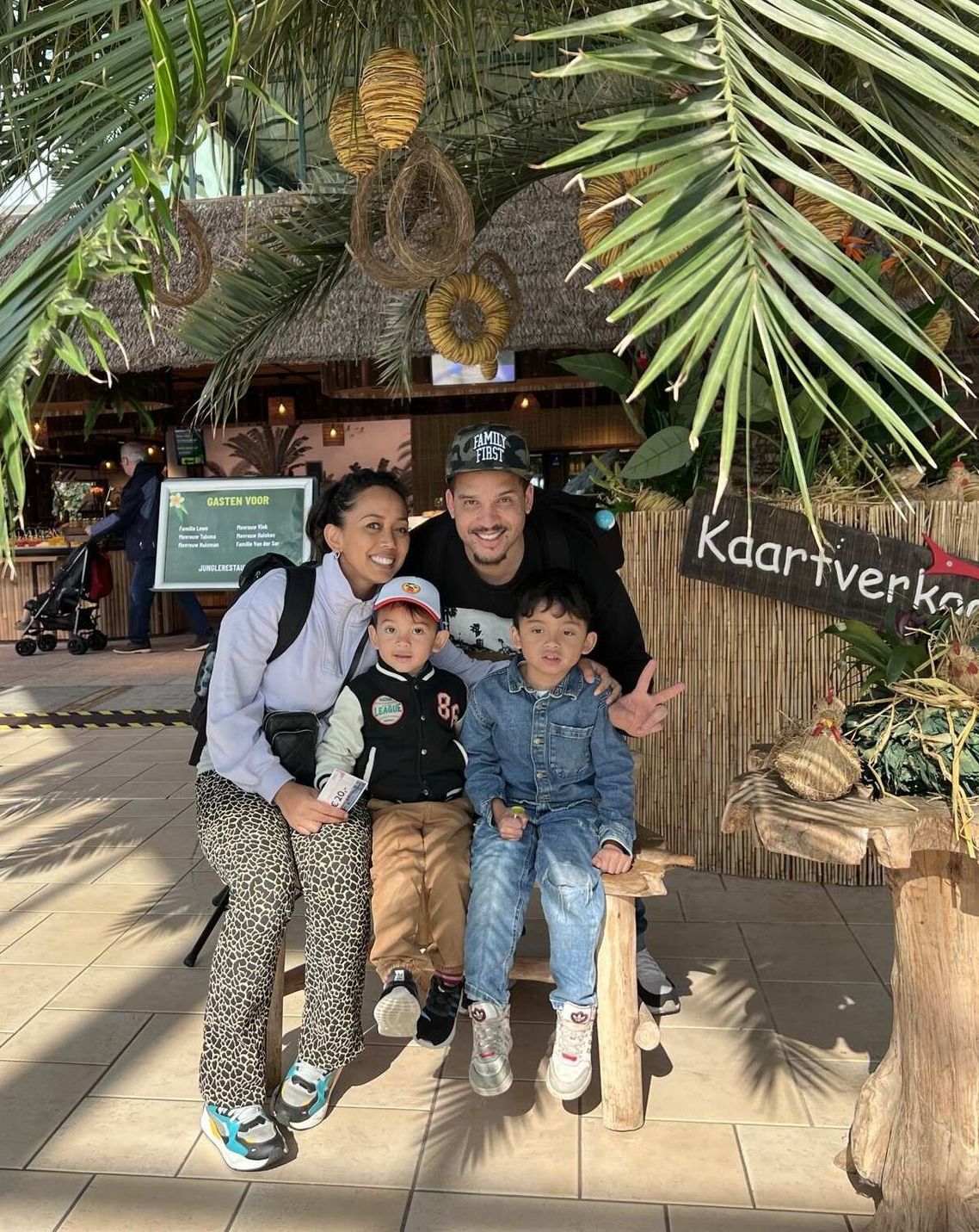 Een gezin poseert voor een foto bij een kiosk met tropisch thema. Palmbladeren hangen boven hun hoofden. Twee lachende kinderen staan vooraan.