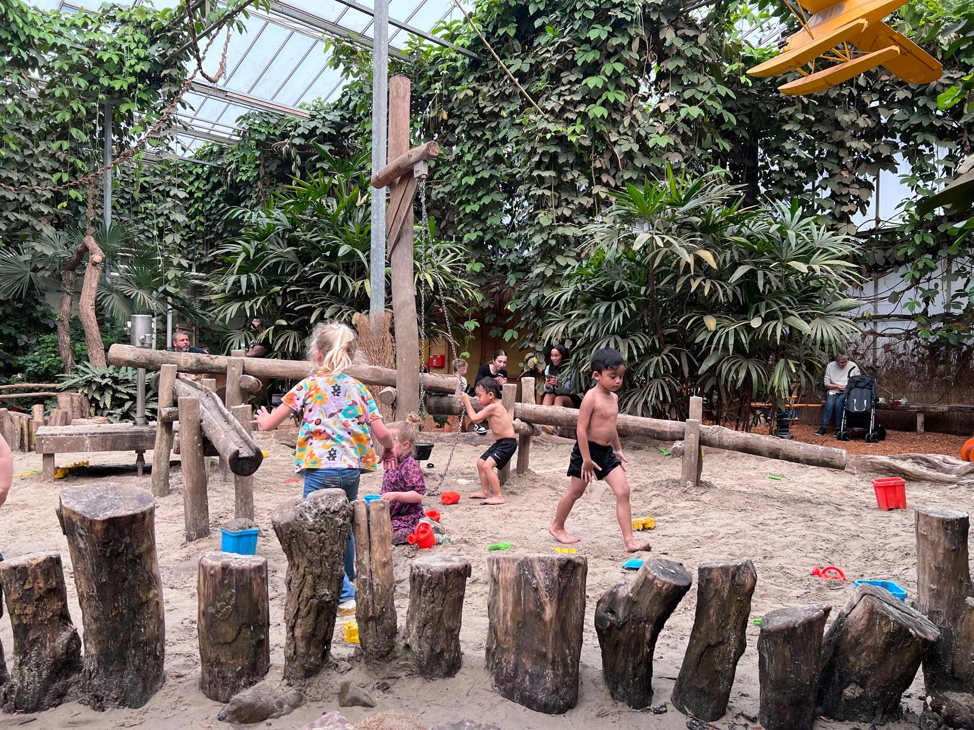 Kinderen spelen in een zandbak, omringd door houten boomstammen en groen.