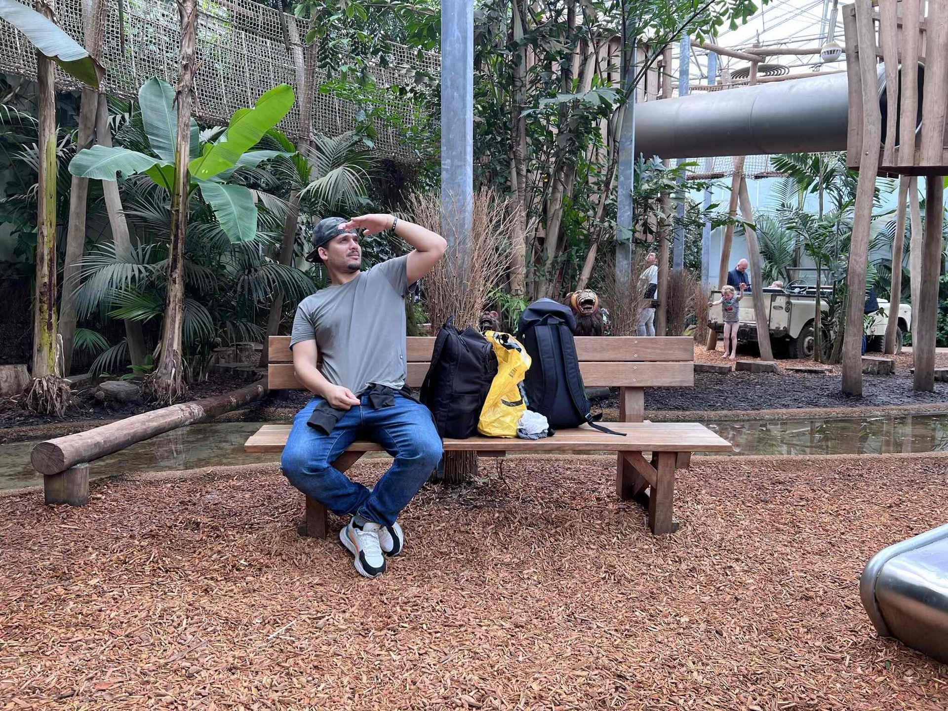 Een man zit met rugzakken op een parkbankje en kijkt omhoog. Hij is omringd door planten en een speeltoestel.