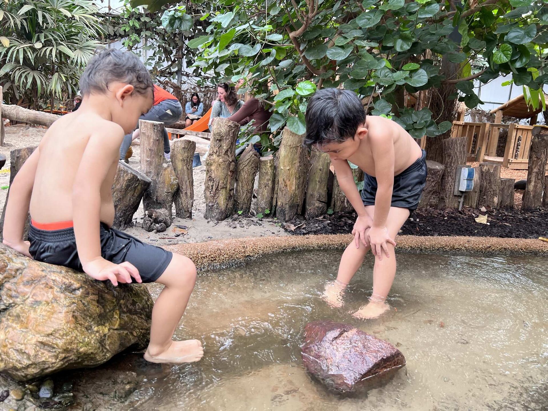 Twee kinderen spelen in een ondiep zwembad. De ene zit op een rots, de andere staat, beiden dragen een zwembroek.
