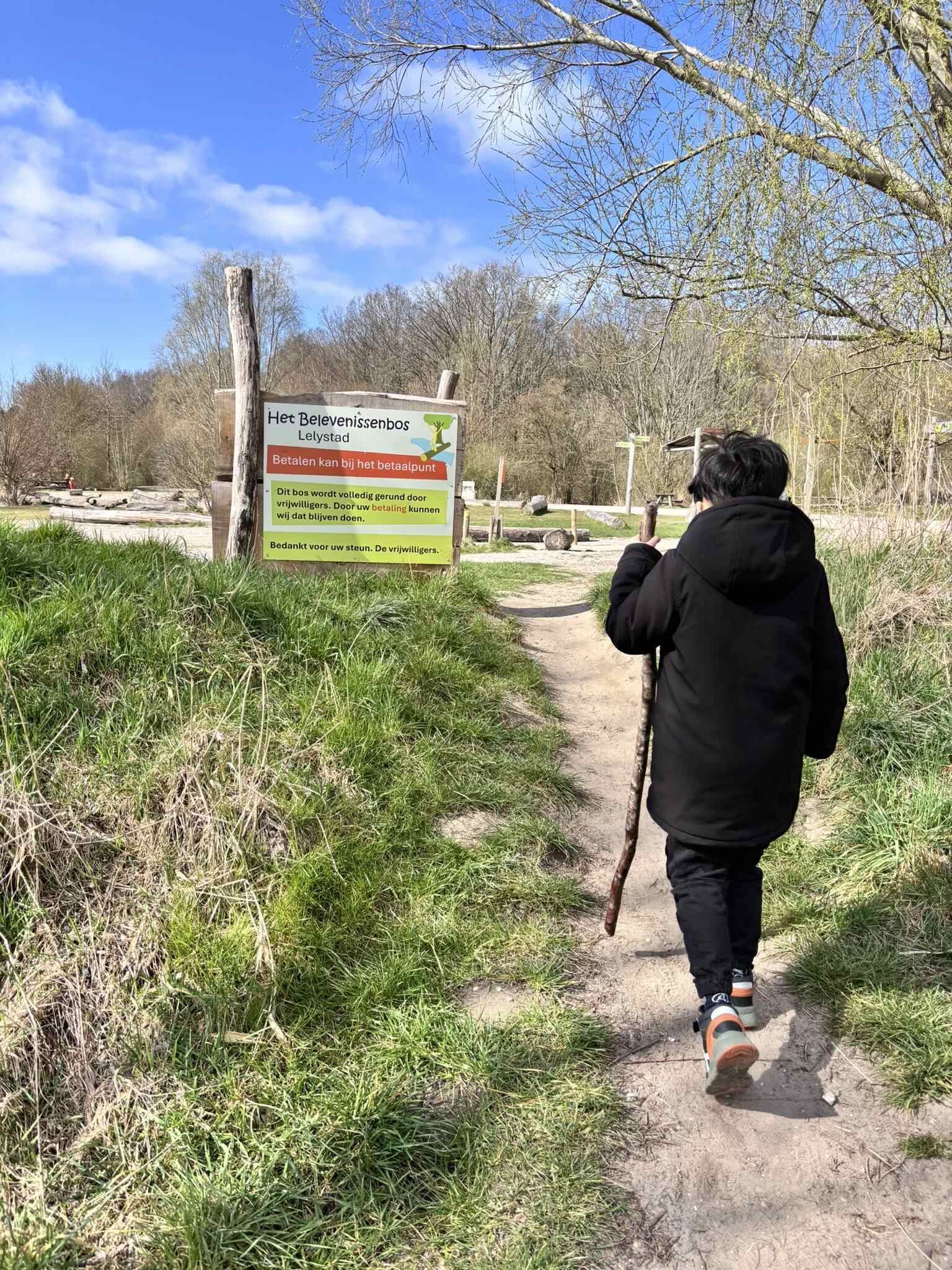 Een persoon loopt met een stok in zijn hand over een zandpad richting een bord in een park.
