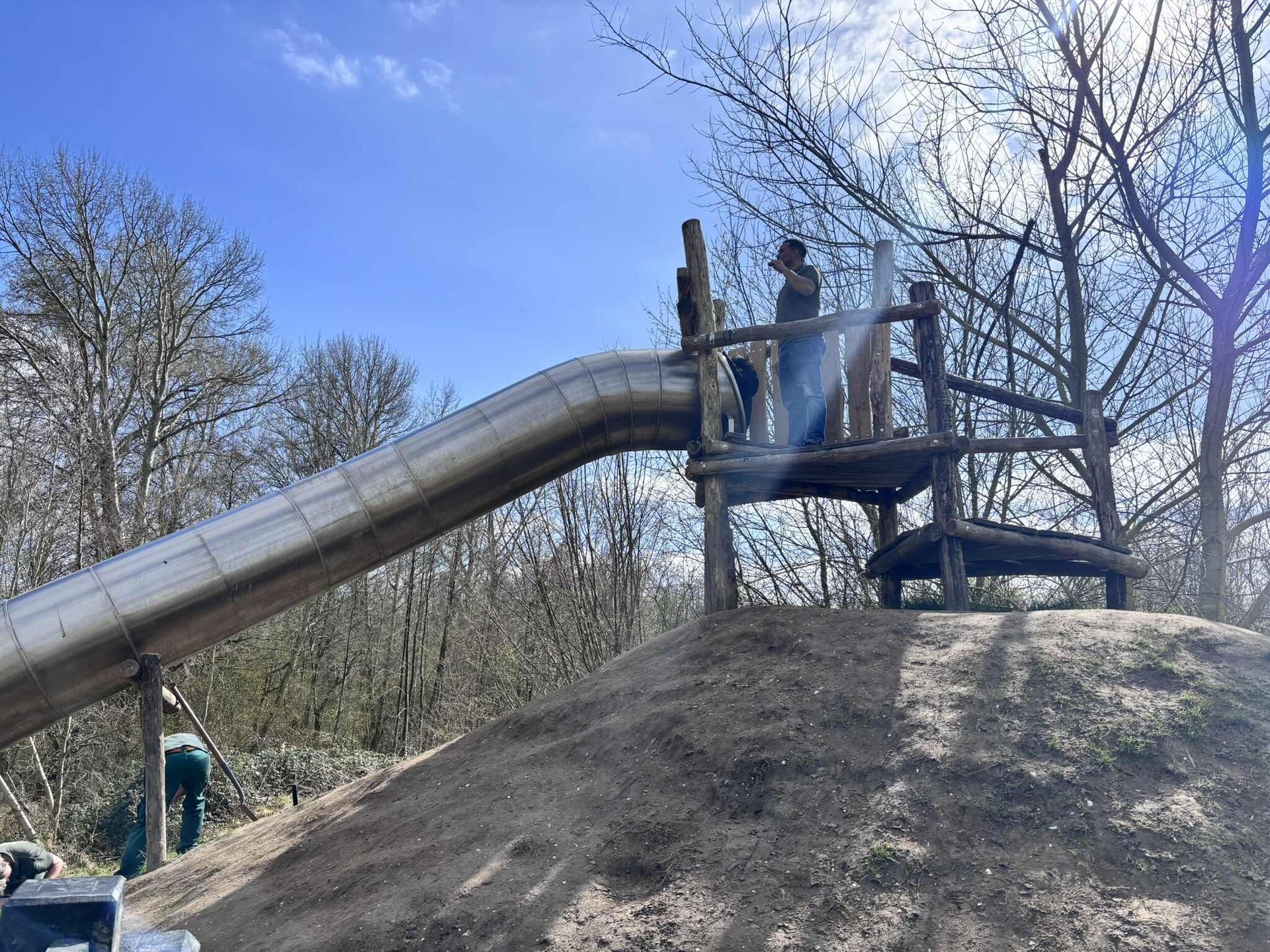 Een metalen glijbaan op een heuvel, met een persoon die op een houten platform bovenaan staat, en bomen op de achtergrond.