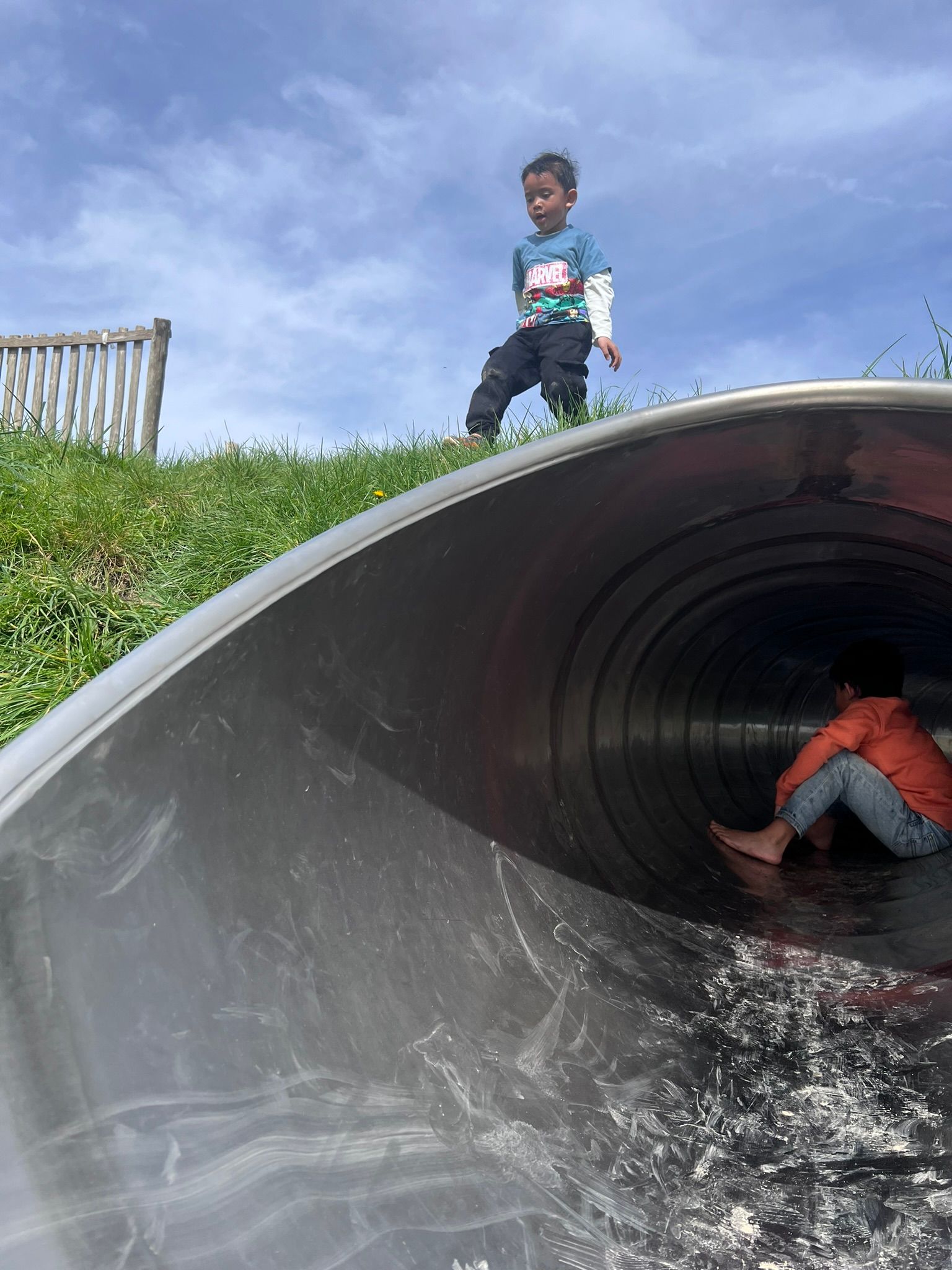 Twee kinderen spelen op een metalen glijbaan in de vorm van een tunnel op een grasheuvel; de ene glijdt naar beneden, de andere staat erboven.