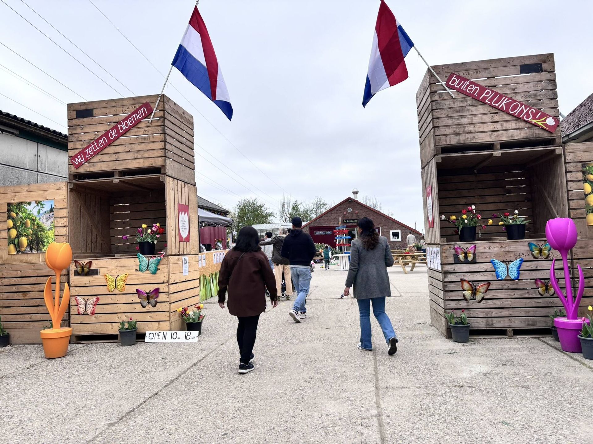 Mensen lopen richting een ingang met houten constructies versierd met bloemen en Nederlandse vlaggen. Bewolkt weer.