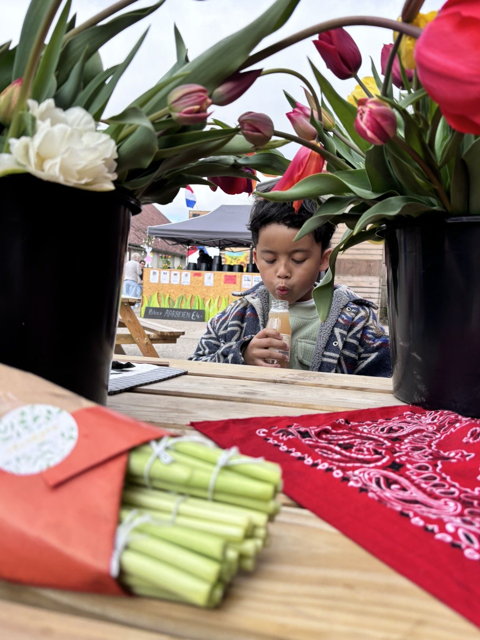 Een jongen nipt aan een drankje aan een tafel met bloemen in potten. Er liggen bosjes citroengras en een rood tafelkleed op de tafel.