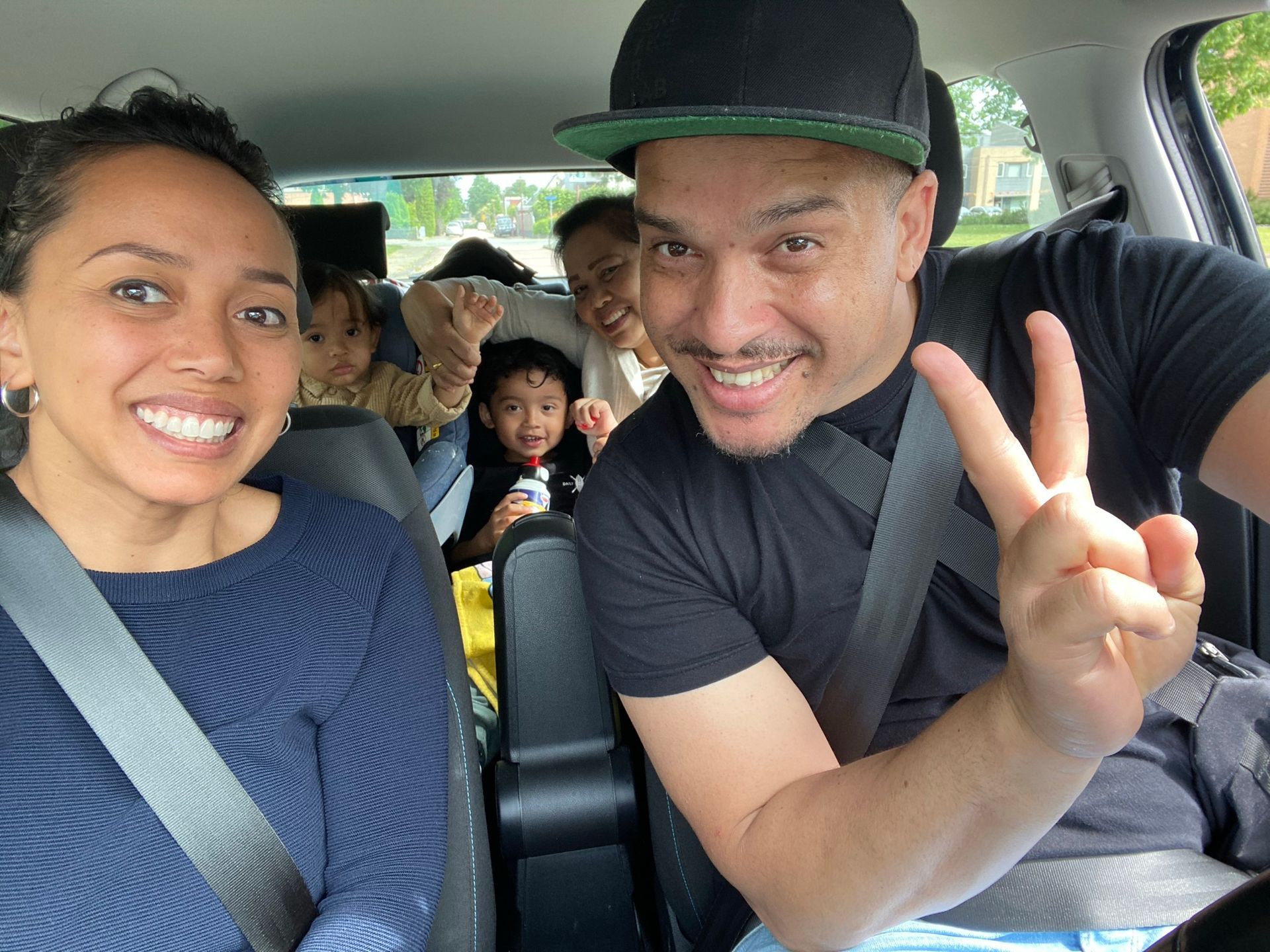 Een gezin zit lachend in een auto. Een man met een hoed maakt een vredesteken. Kinderen zitten achterin.