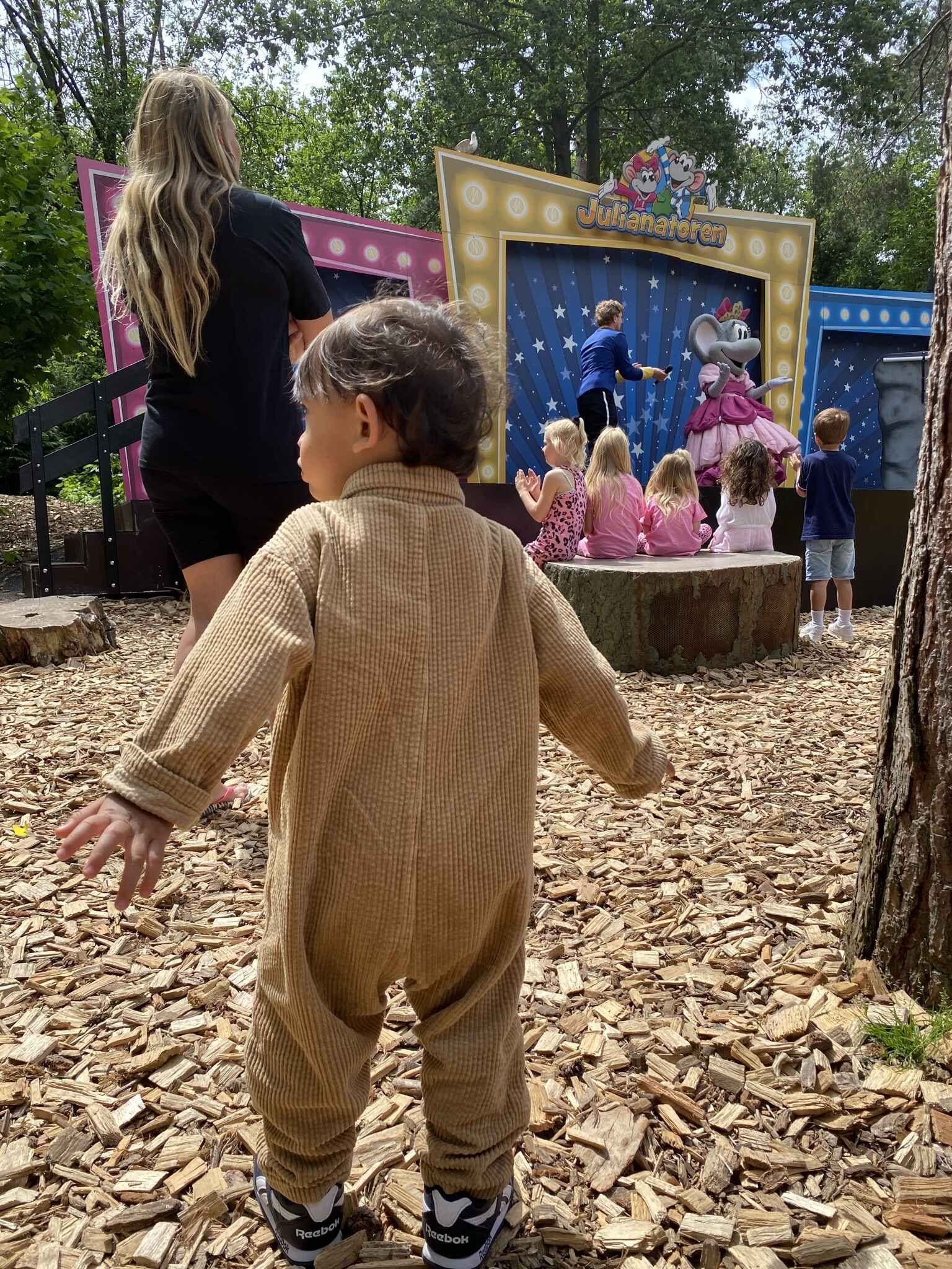 Een kind in een bruine overall met uitgestrekte armen kijkt naar een theatervoorstelling in de buitenlucht.