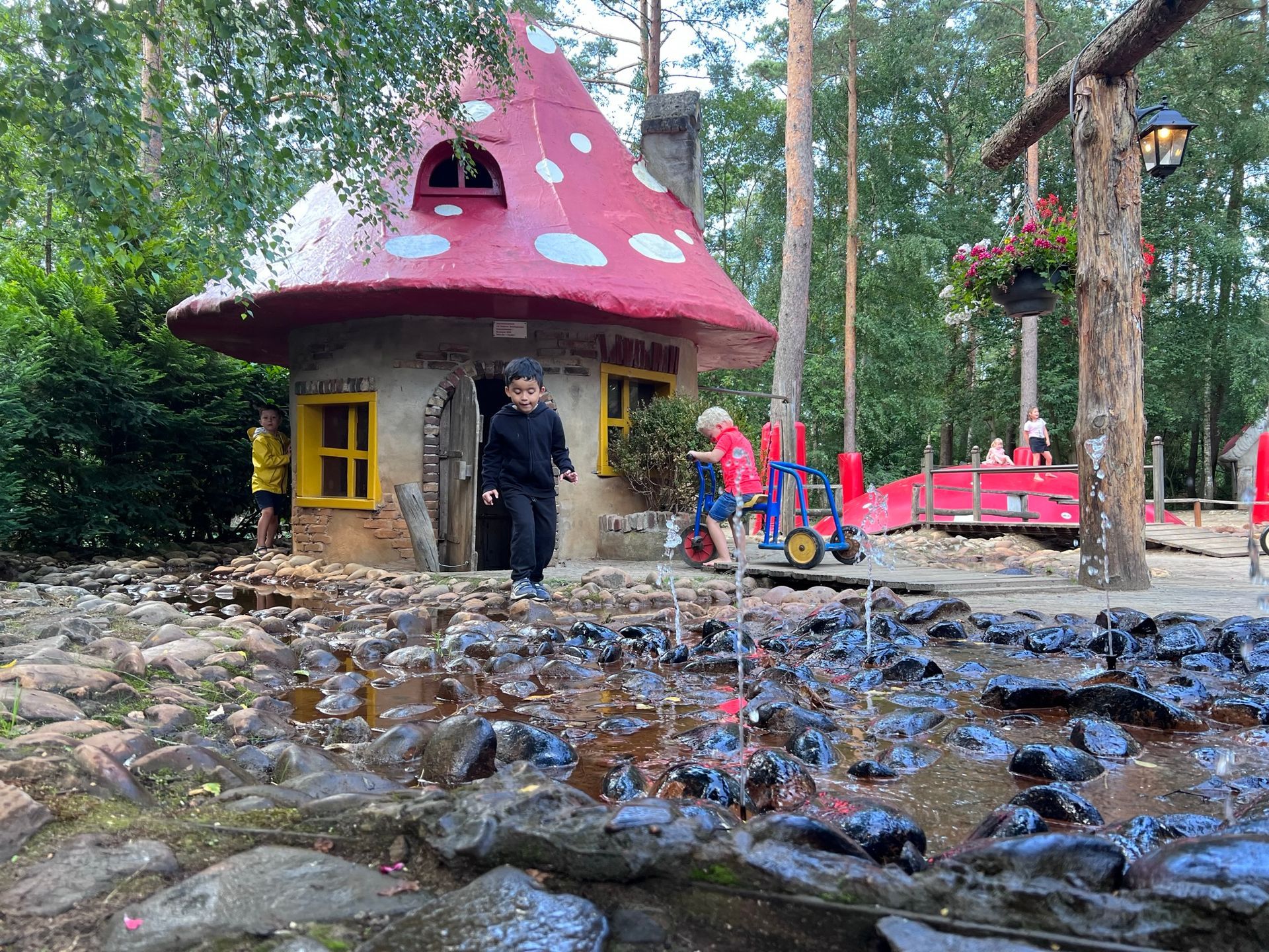 Een paddenstoelvormig speelhuisje met kinderen. Een kind loopt naar de camera toe. Buiten in een park.