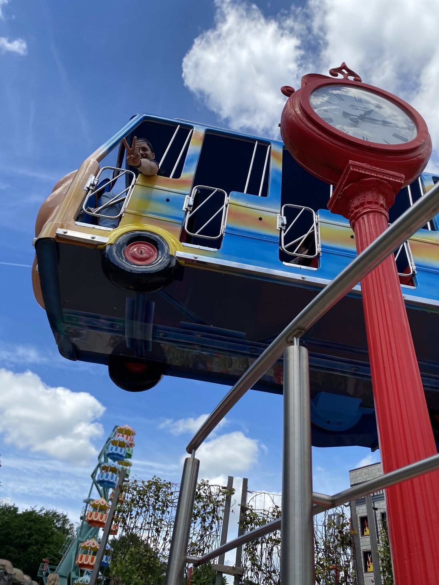 Een attractie in een pretpark met een blauwe, busvormige wagen in de lucht, een rode klokkentoren en een reuzenrad.
