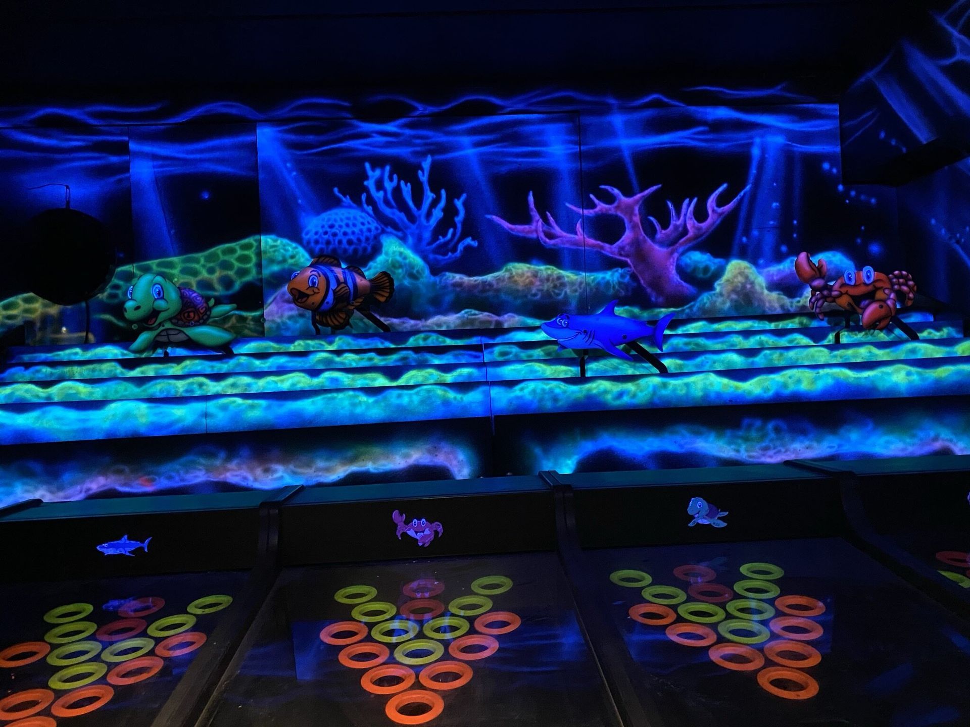 Een kamer met een oceaan-thema, verlicht door blacklight, met oplichtend koraal, vissen en oranje/groene doelen in verschillende niveaus voor een spel.