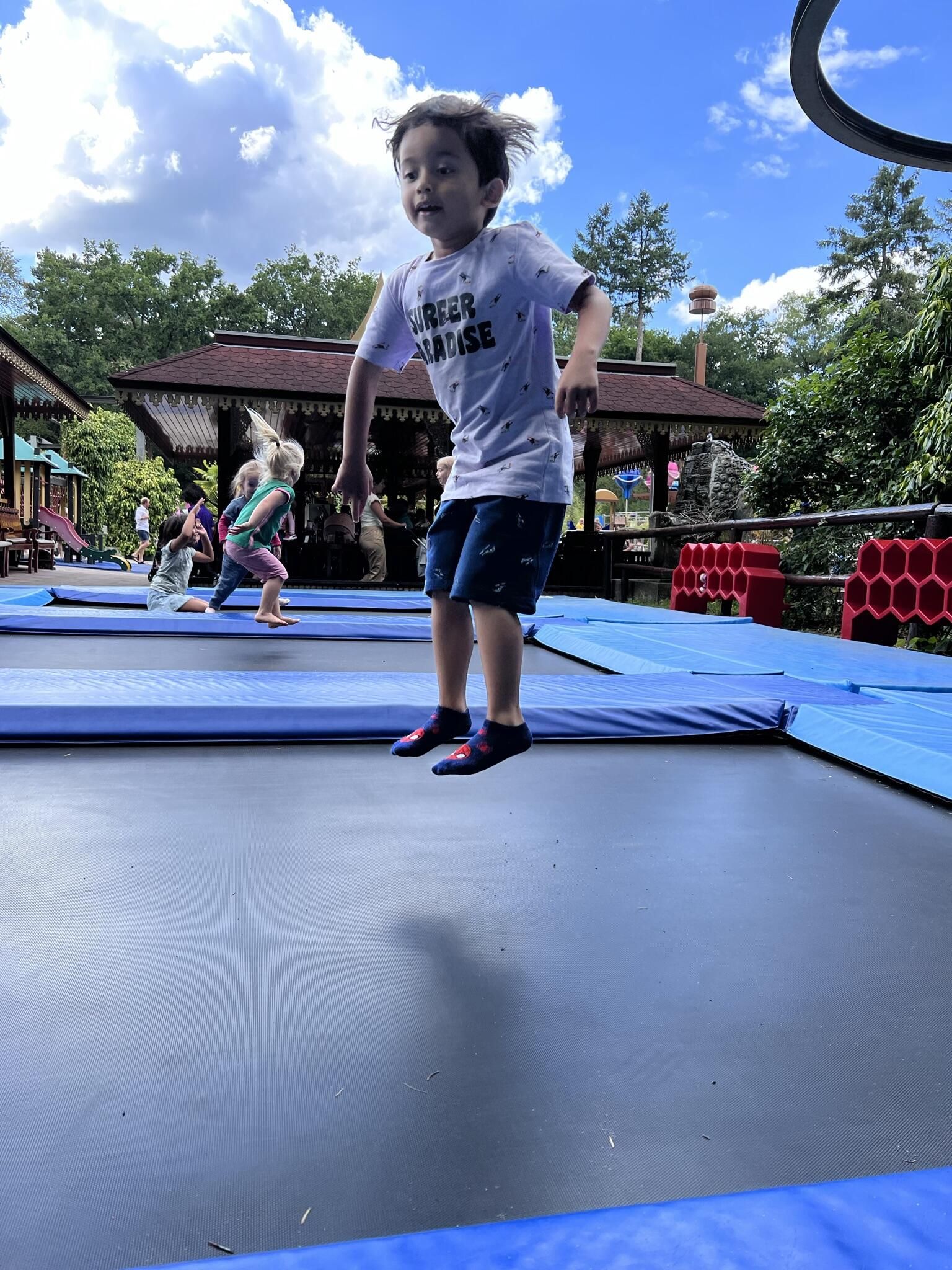 Een jongen springt op een trampoline onder een blauwe hemel, andere kinderen spelen op de achtergrond.