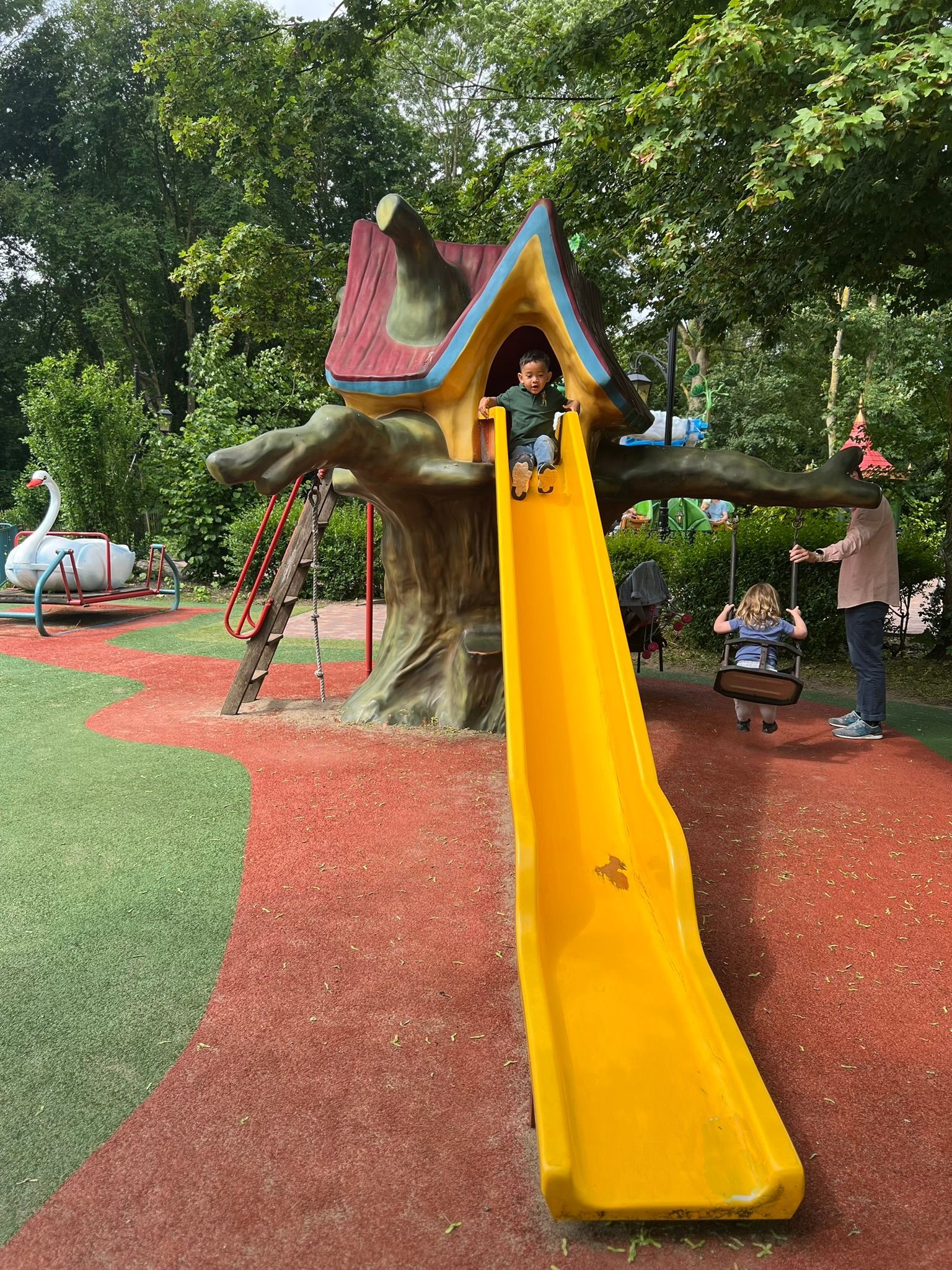 Een kind glijdt van een gele glijbaan op een speeltuin met een boomhut.