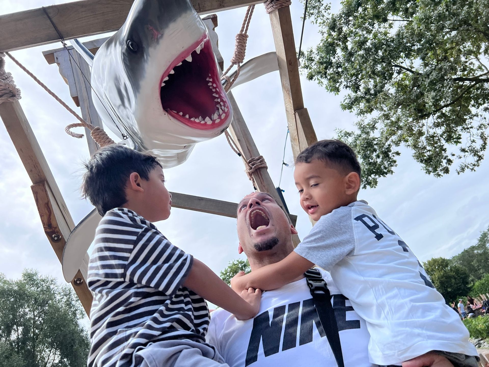 Een man met open mond, terwijl twee kinderen aan zijn shirt trekken, staat onder een haaieninstallatie in een pretpark.
