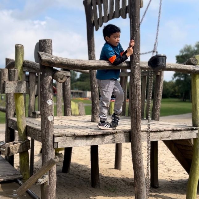 Kind in een turquoise shirt en grijze broek staat op een houten speeltoestel en houdt zich vast aan een ketting.