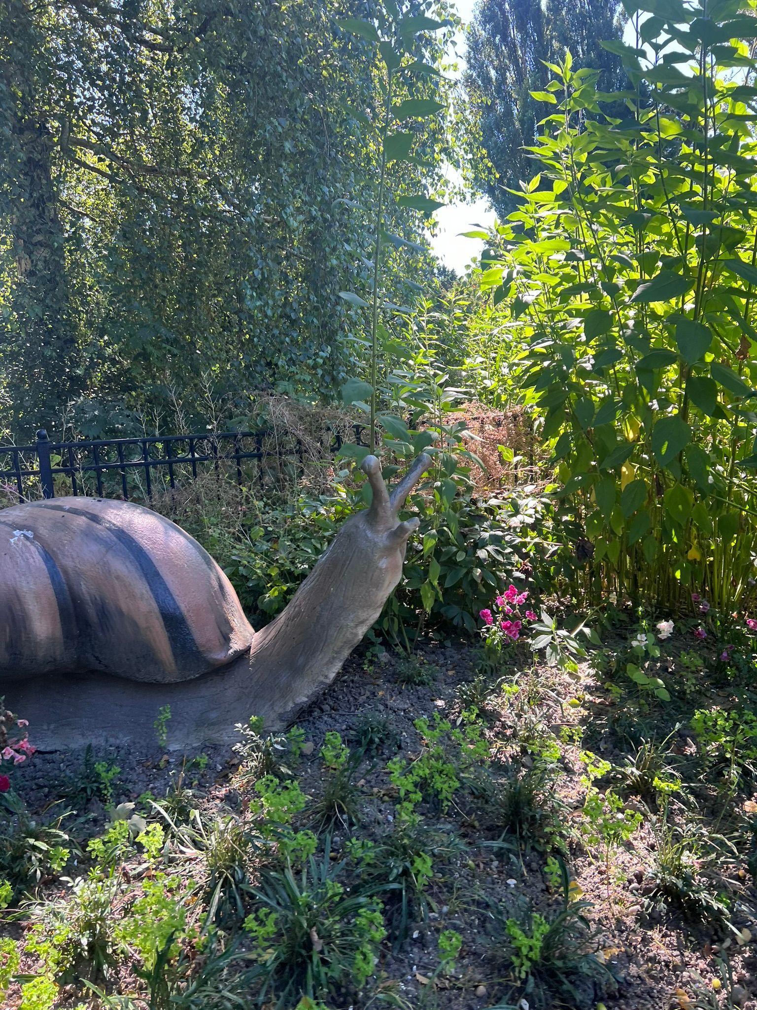 Een groot slakkenbeeld in een tuin, dat zijn voelsprieten omhoog uitstrekt, naast begroeiing en een hek.