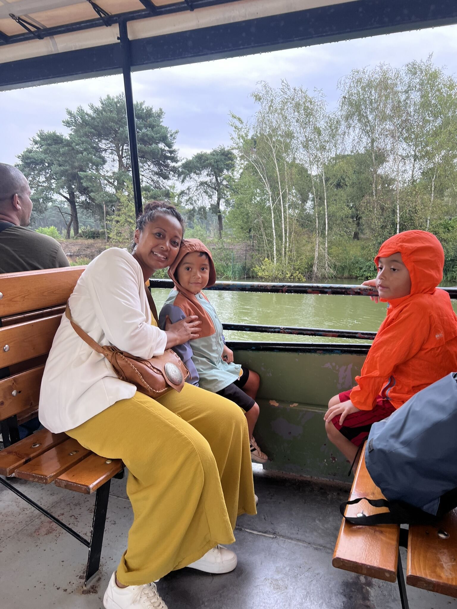 Vrouw en twee kinderen in een boot, lachend. Bomen en water op de achtergrond. Bewolkte lucht.