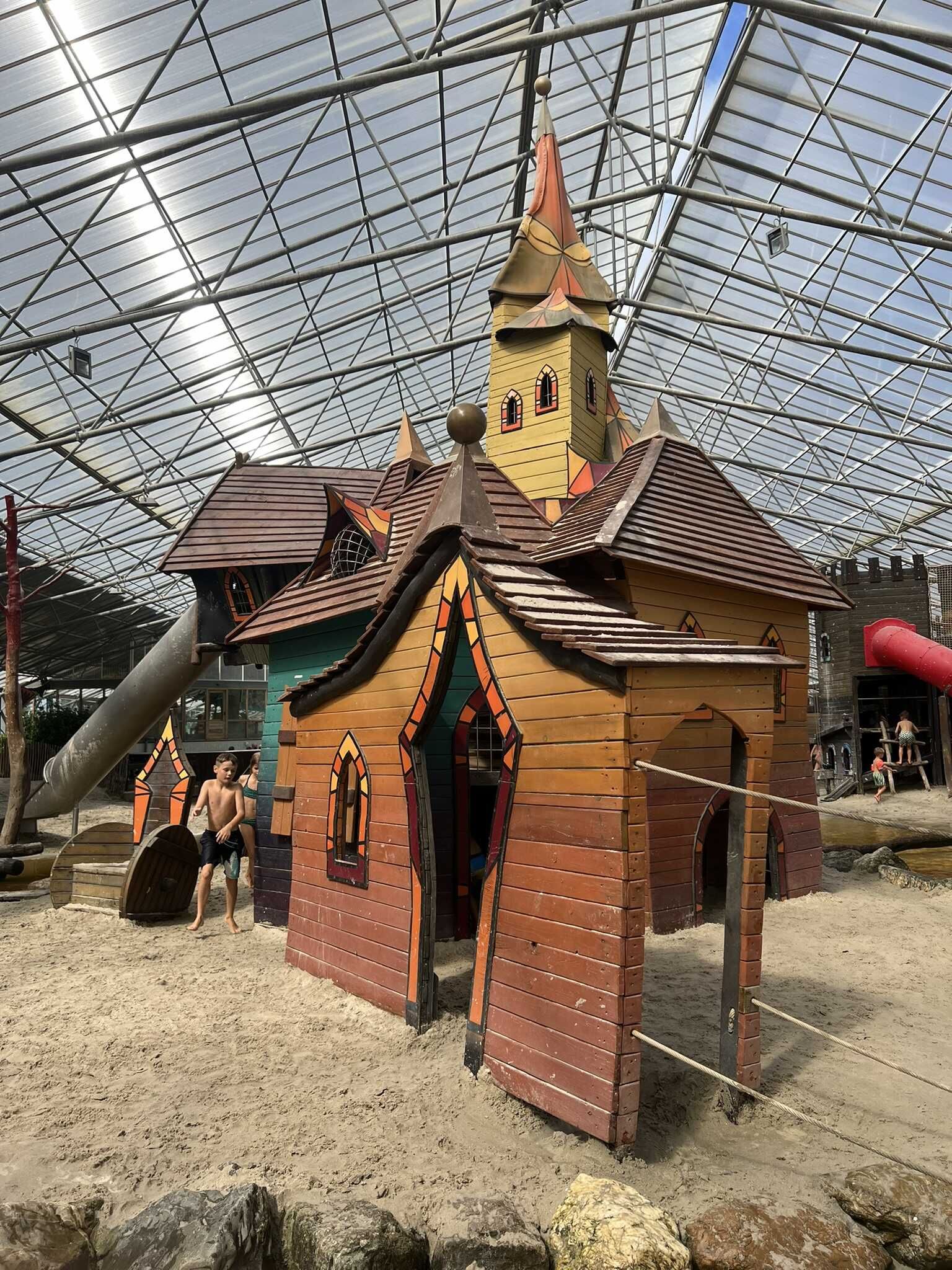 Speelstructuur met torens, glijbanen en zand in een binnenspeeltuin.