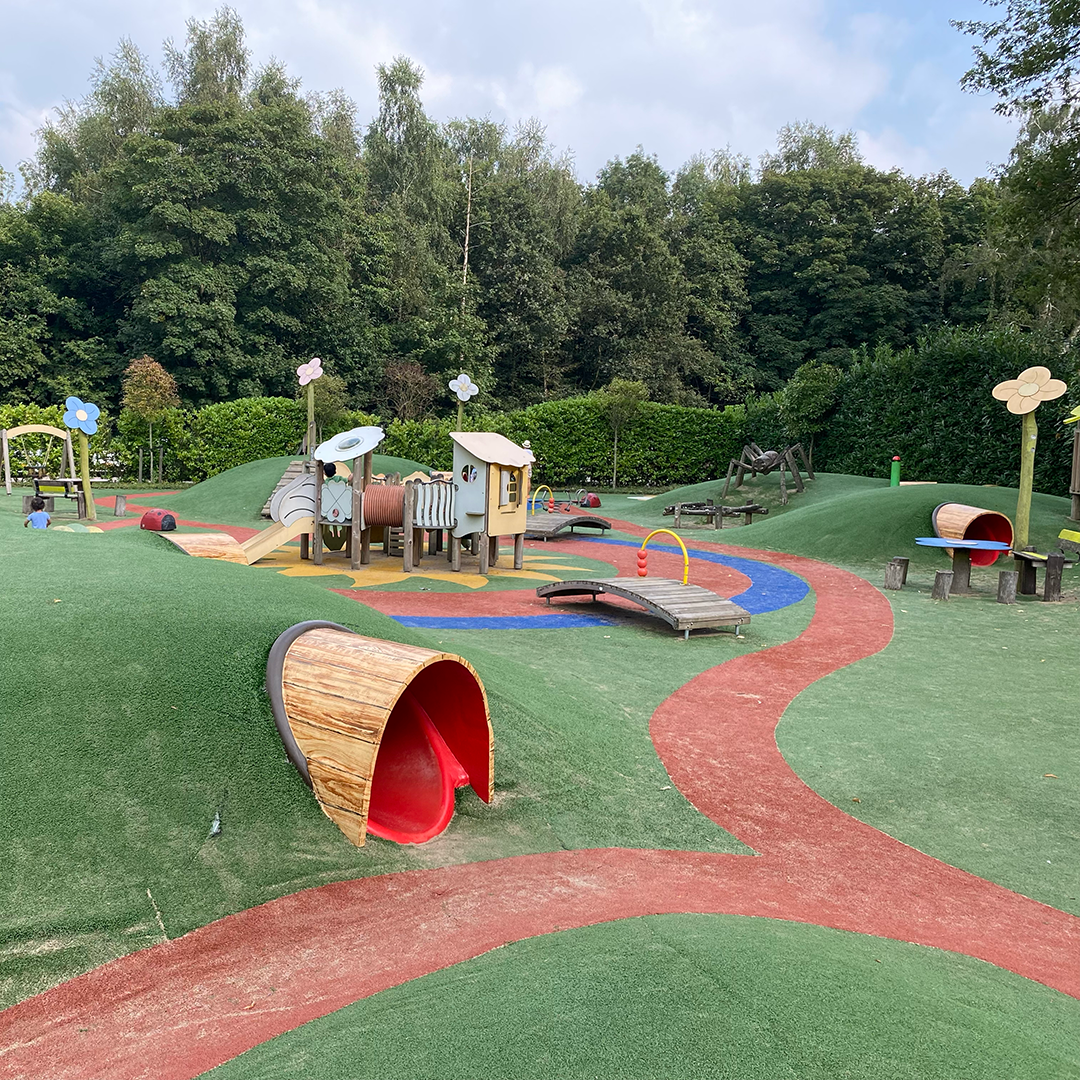 Speeltuin met groen gras, rode paden en houten speeltoestellen. Bos op de achtergrond.