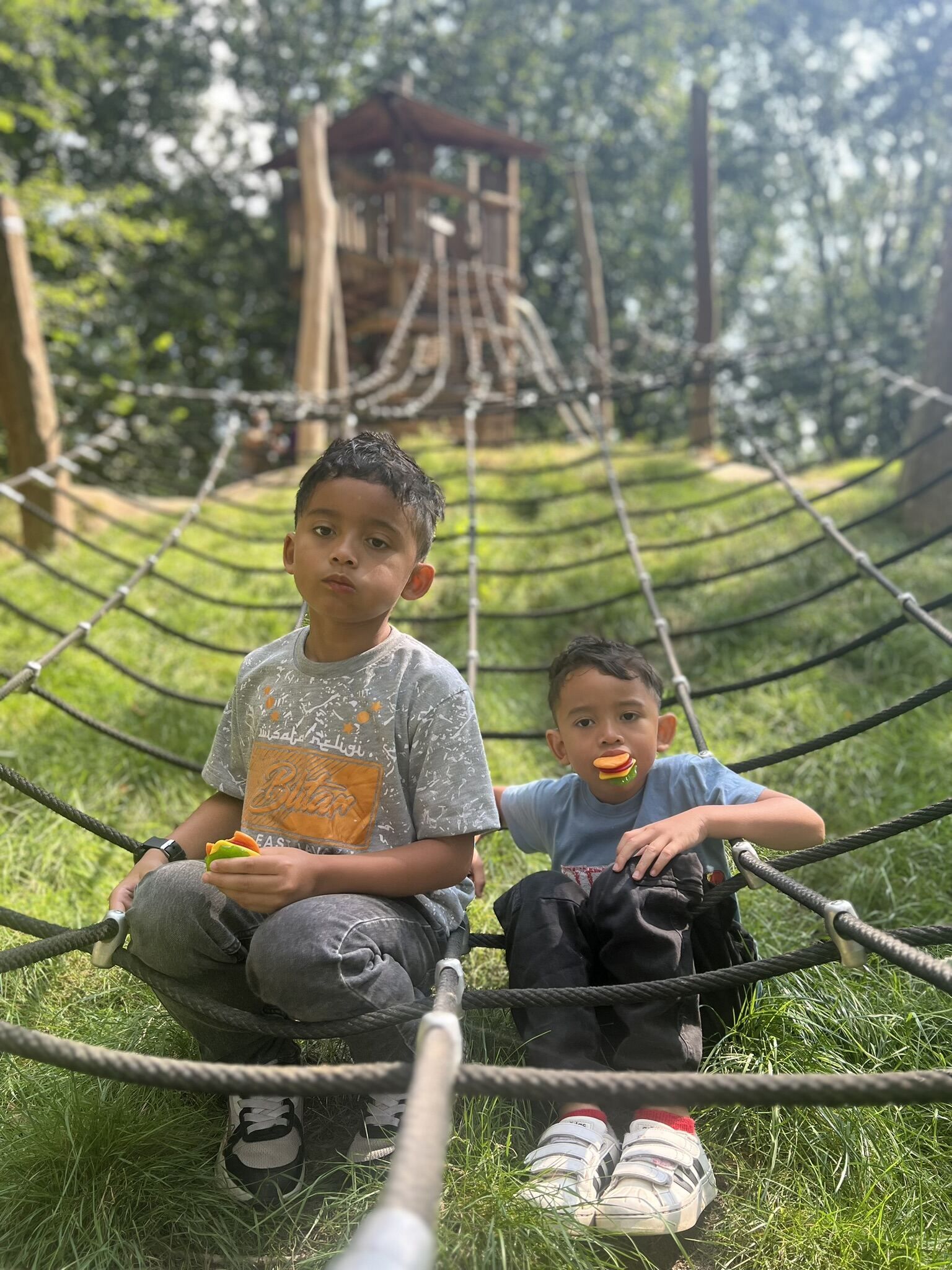 Twee kinderen zitten op een touwnet in een park en houden snoepjes vast. Op de achtergrond is een houten speeltoestel te zien.