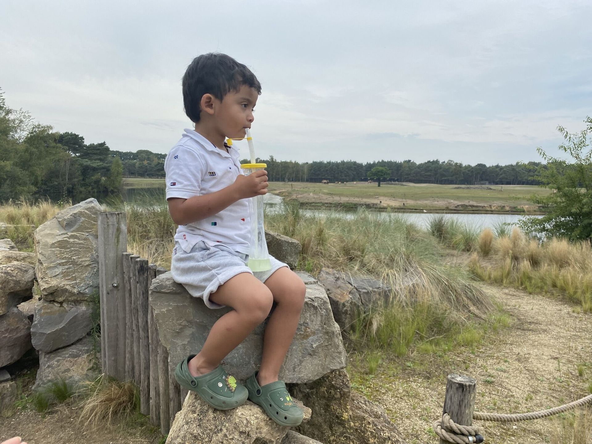 Een jong kind drinkt iets terwijl het op een rots zit, vlakbij een waterpartij en bomen.