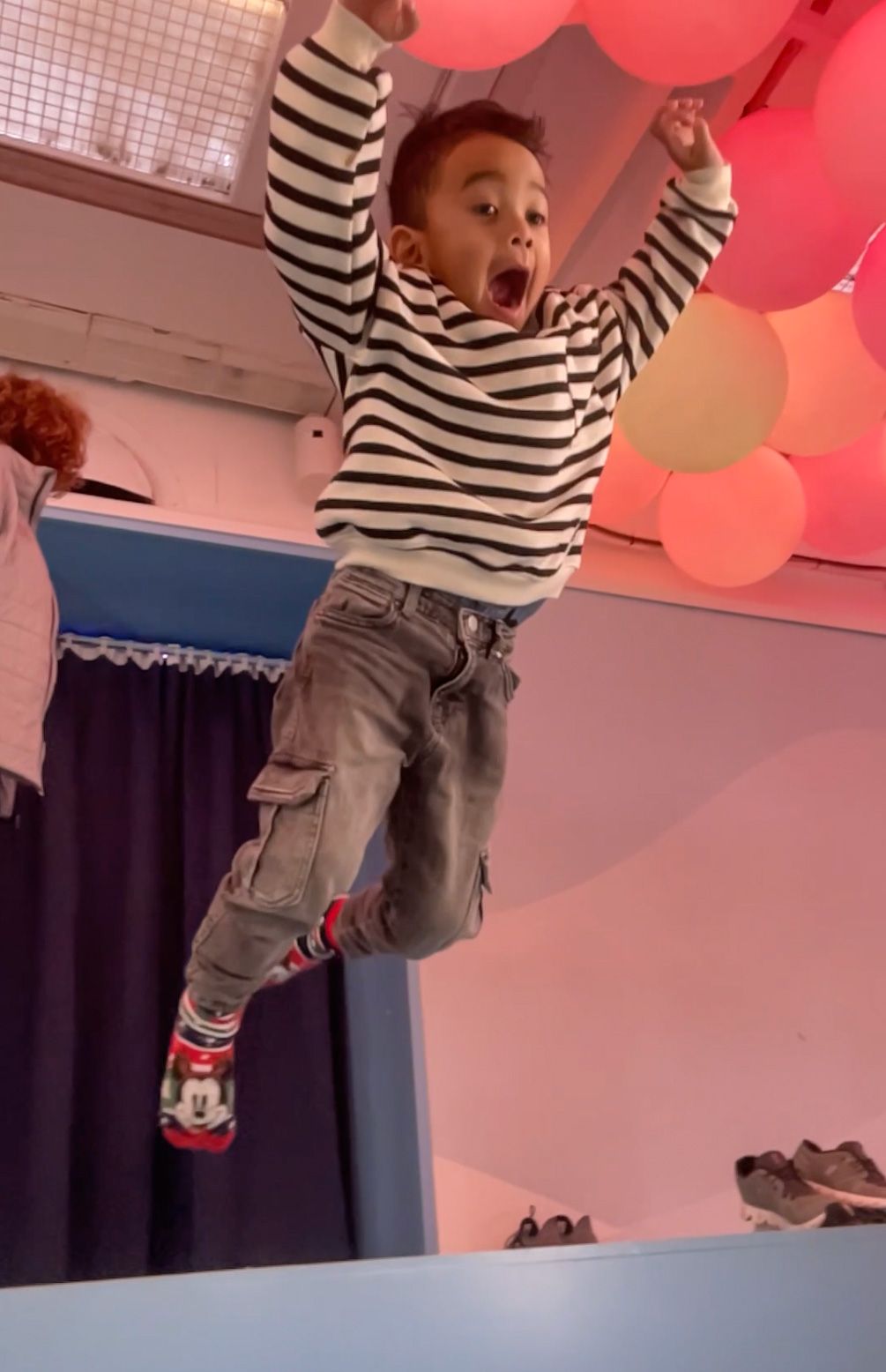Jongen springt met opgeheven armen en een opgewonden uitdrukking. Gestreepte trui, cargobroek, roze ballonnen op de achtergrond.