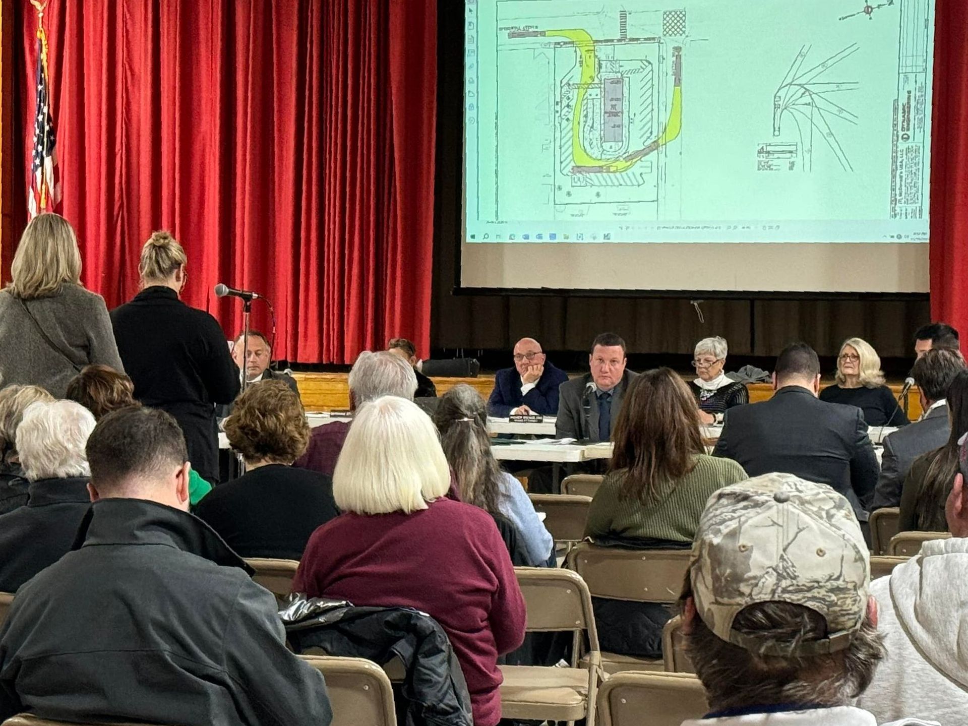 A public meeting with attendees, a panel at a table, and a projection screen with a map.