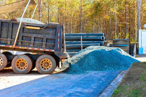 Dump truck unloading blue gravel on a paved surface near trees and stacked pipes.
