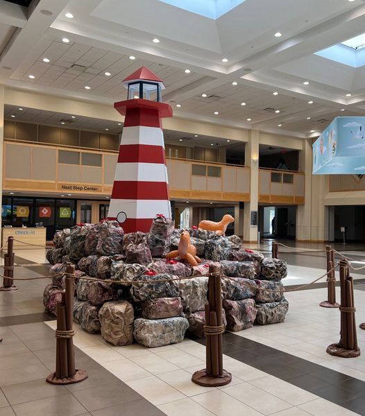 Lighthouse sculpture with red and white stripes, surrounded by rocks and seal statues, in a large interior space.