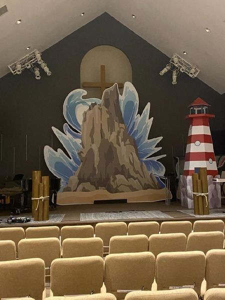 Stage set with a mountain, waves, lighthouse, and cross. Beige seating faces the stage.