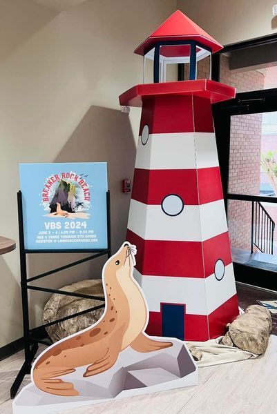 Cardboard lighthouse and seal cutout for VBS 2024, with a sign. Red, white, and blue colors, indoor setting.