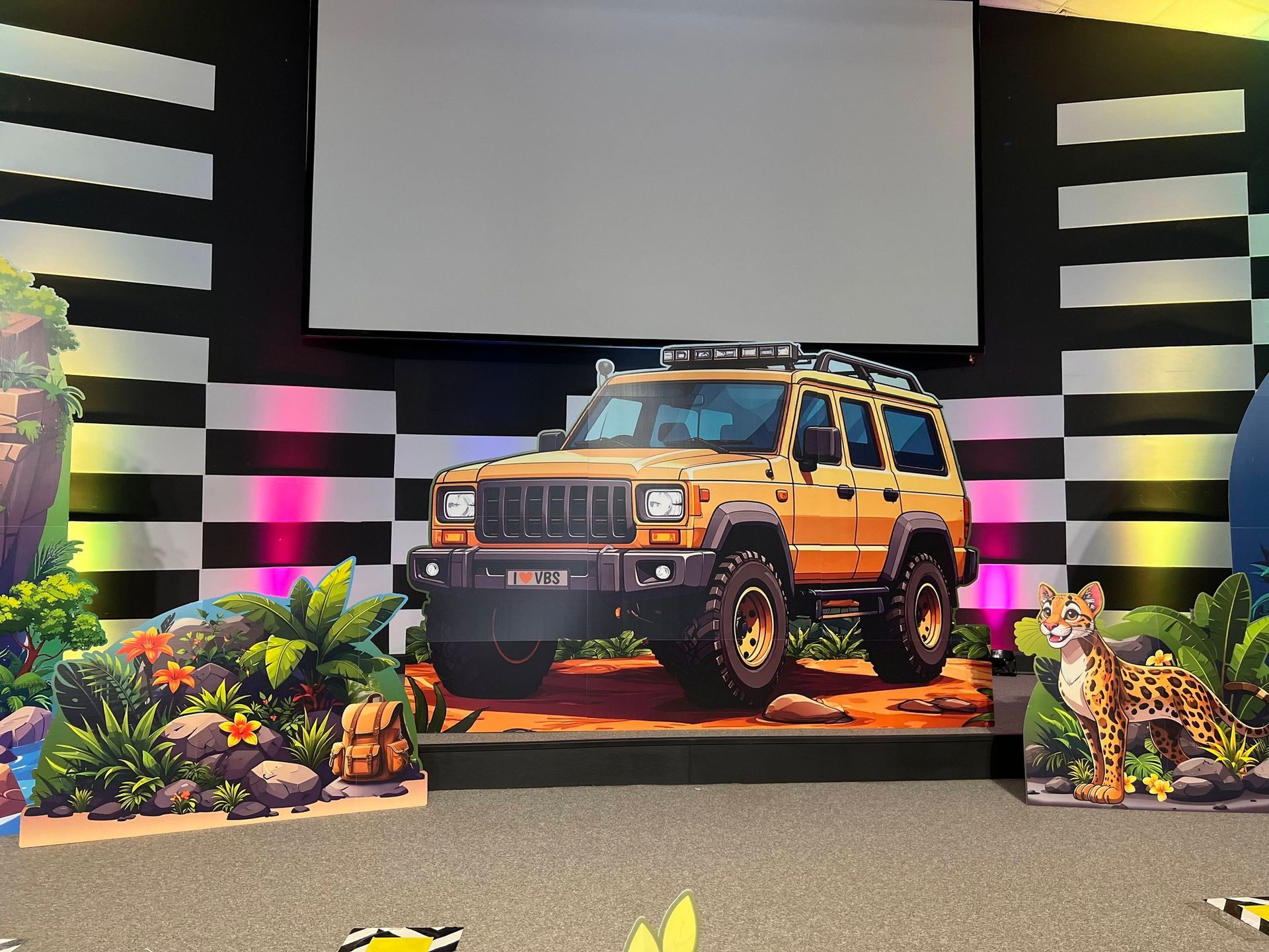 A stage set with a cardboard cutout of a yellow Jeep, jungle-themed decorations, and a projector screen.