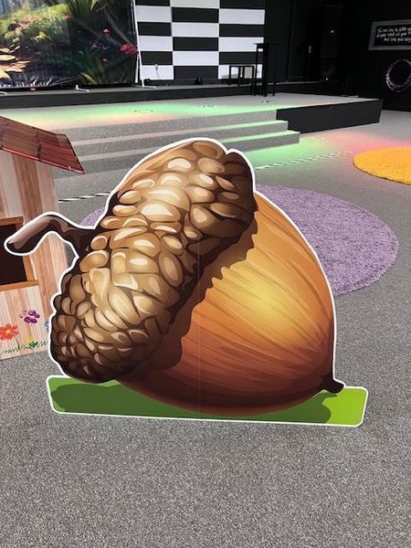 Large cardboard cutout of an acorn on a green base, set in a playroom with a checkerboard backdrop.